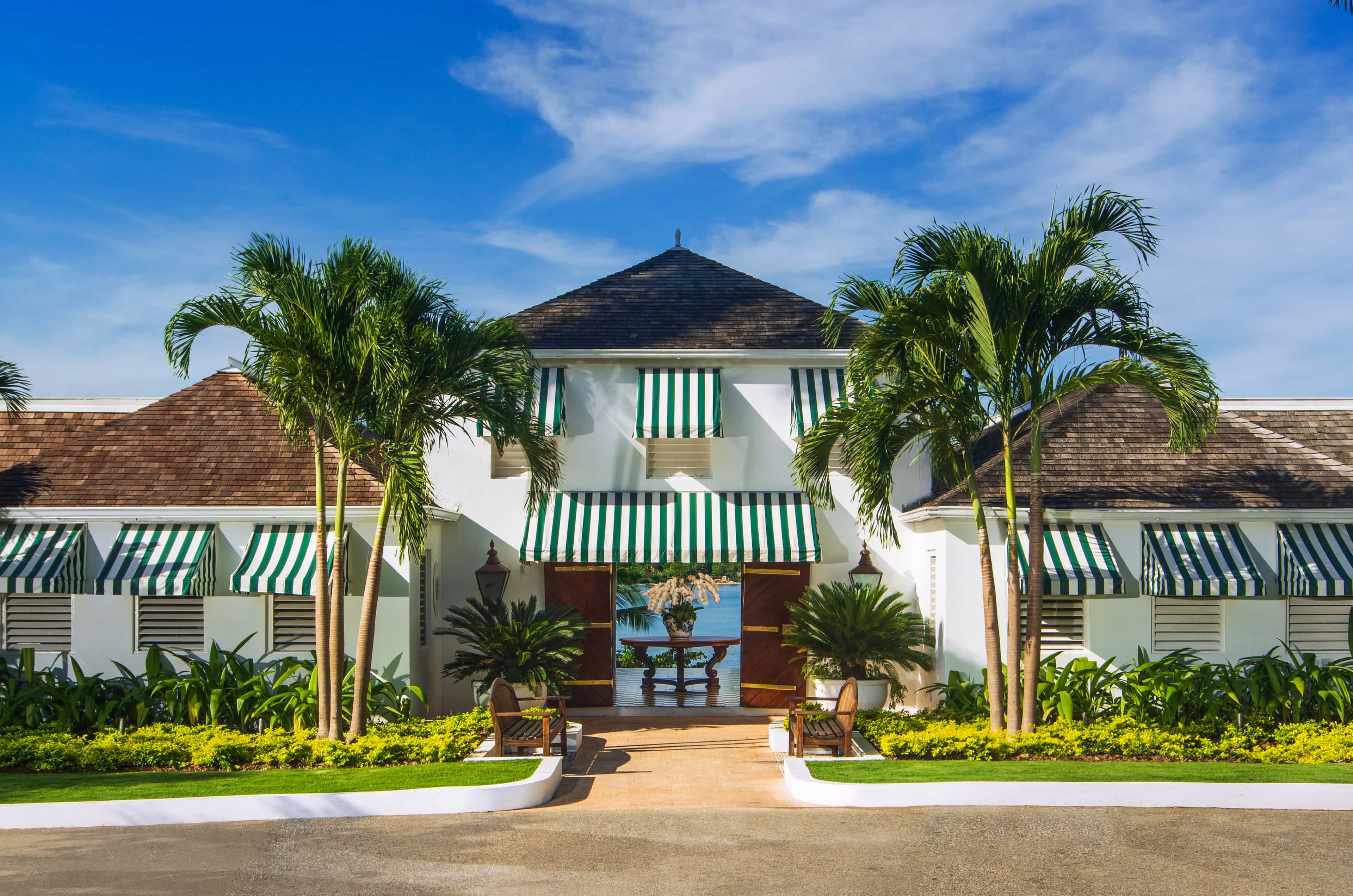 The entrance of a white hotel building with green and white awning and a sea view in the background.