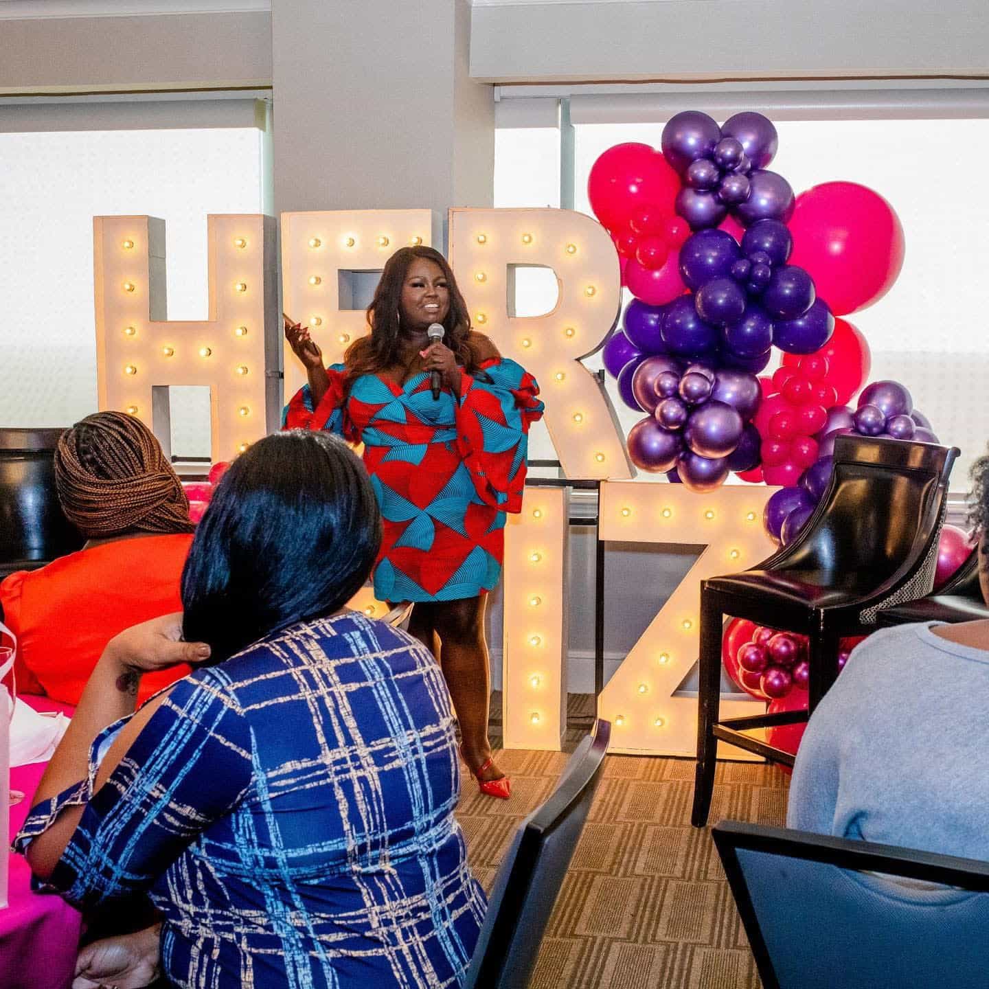 woman in a blue and red dress speaking to a group of people