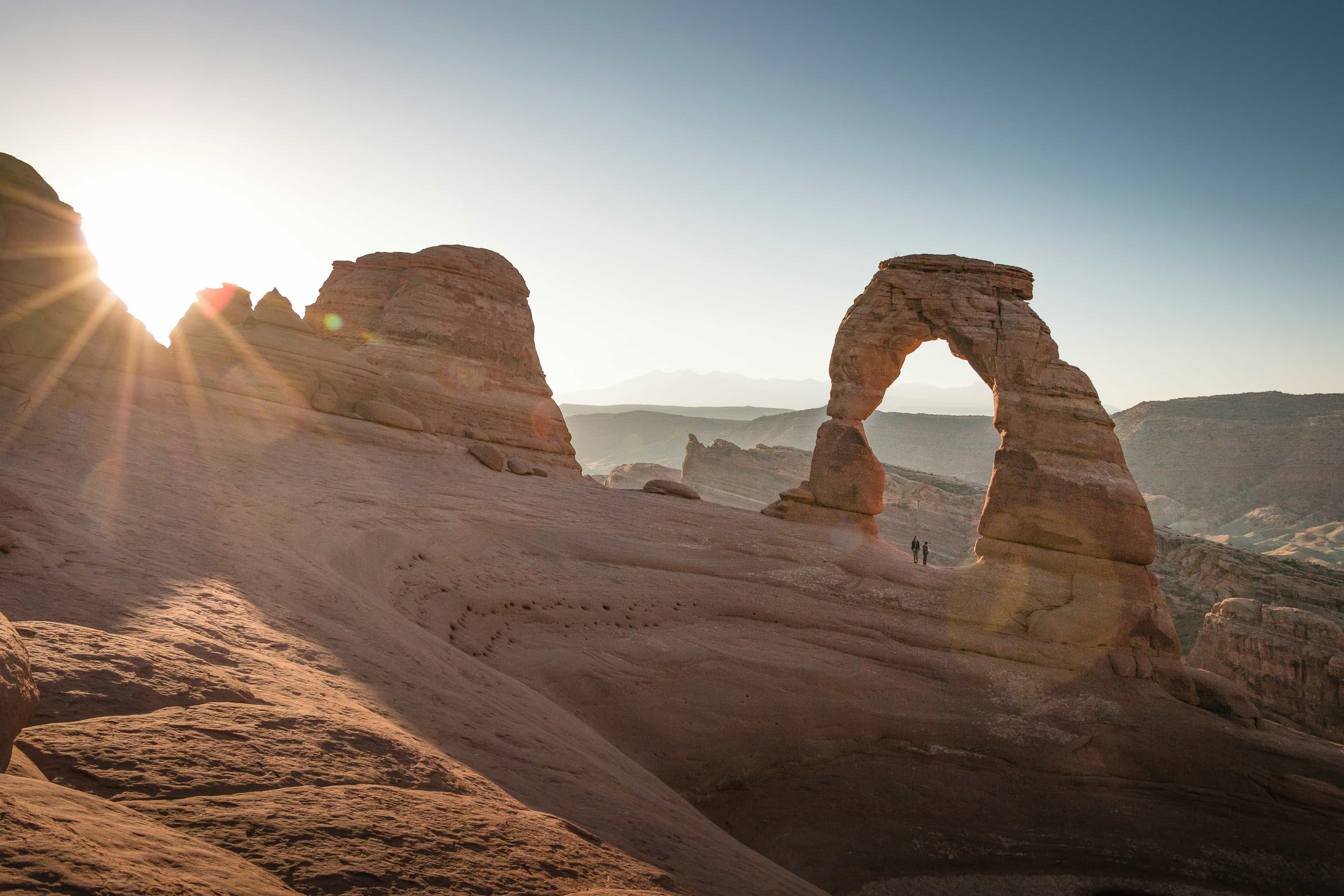 A landscape view of a desert landscape with tan rocks and an arched rock with the sun shining just above the top of the rocks