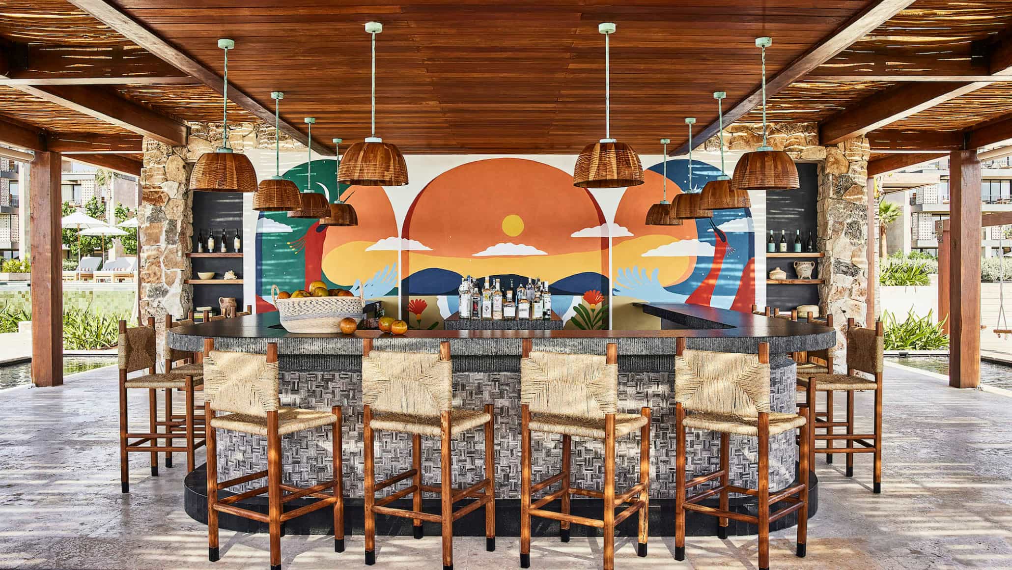 A colorful and modern bar area of a hotel with wooden bar stools and hanging lamps with a mural.