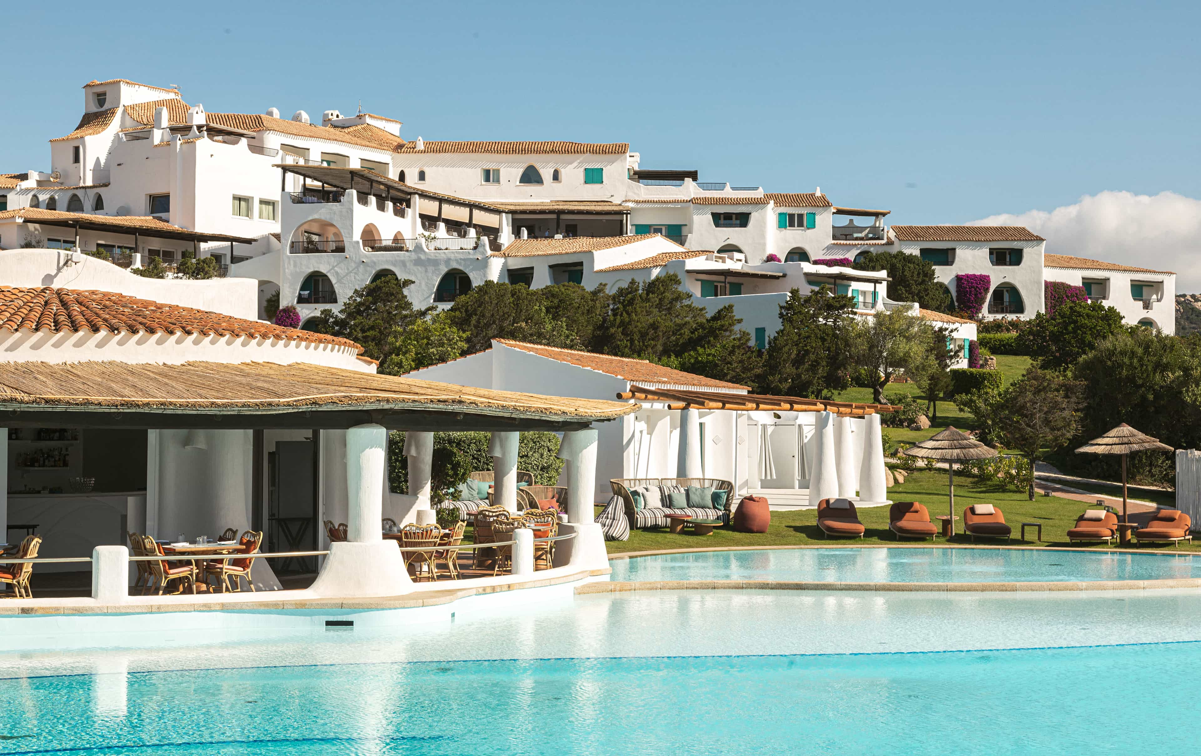 A hotel pool area with cabanas, a restaurant and whitewashed buildings on a hillside behind