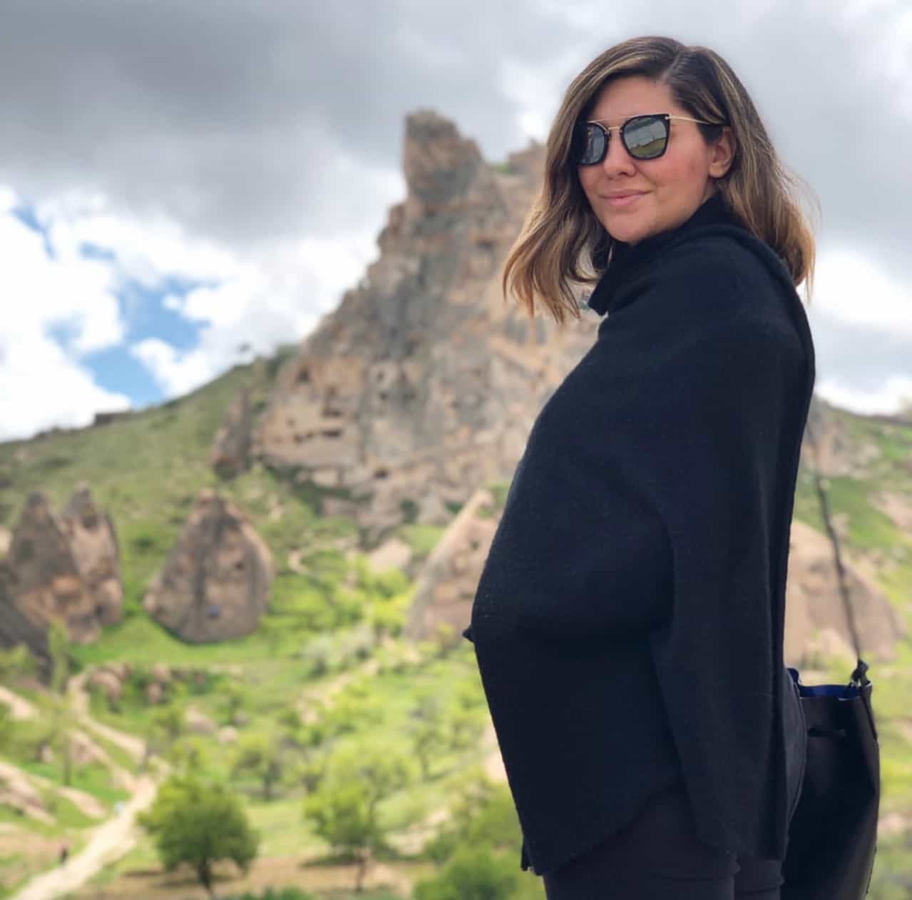 Alejandra in a black jacket and sunglasses posing in front of a mountain landscape on a cloudy day