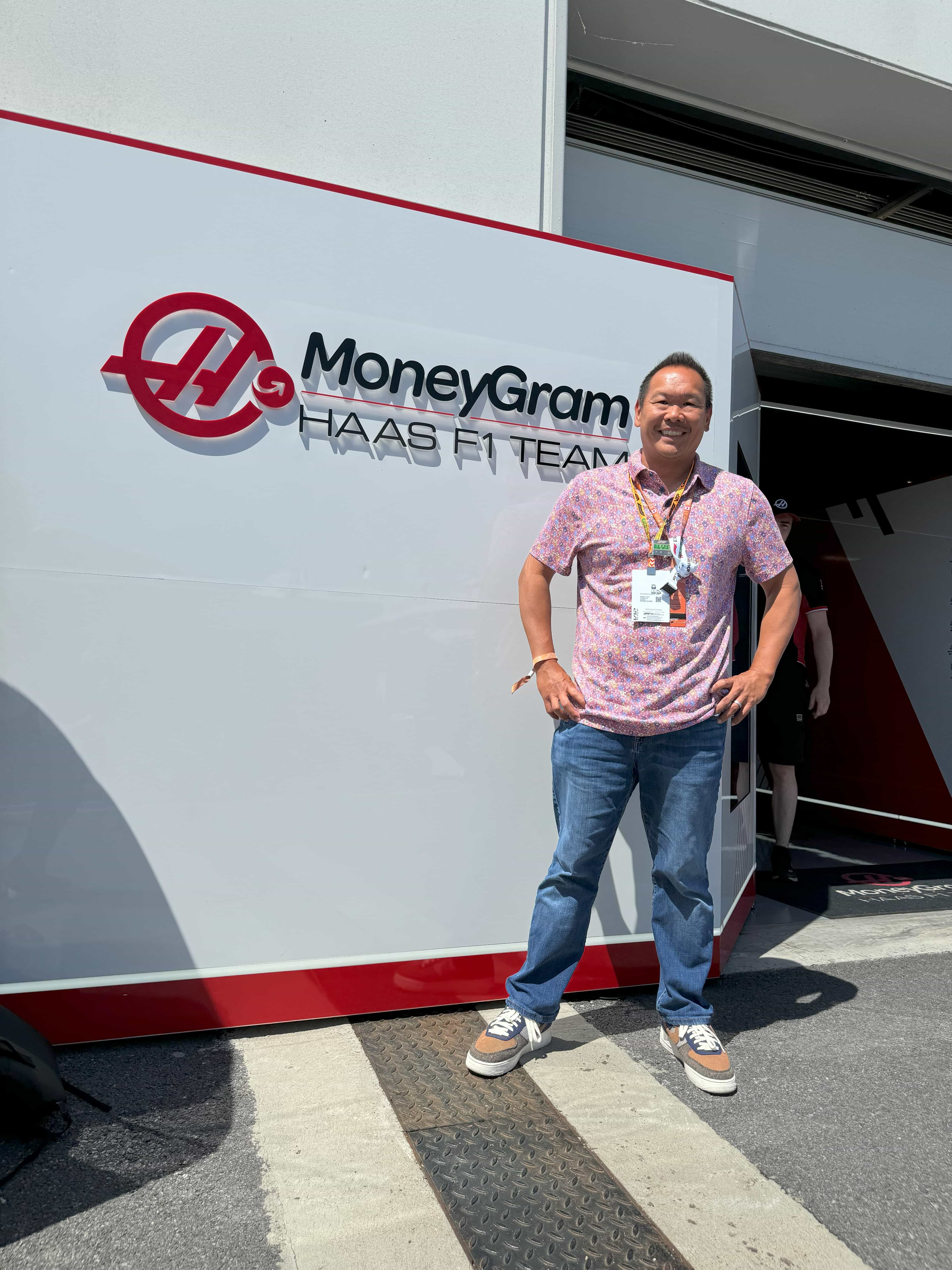 Robert in a pink shirt and jeans standing in front of a white wall with a sign reading “MONEYGRAM” outdoors on a sunny day