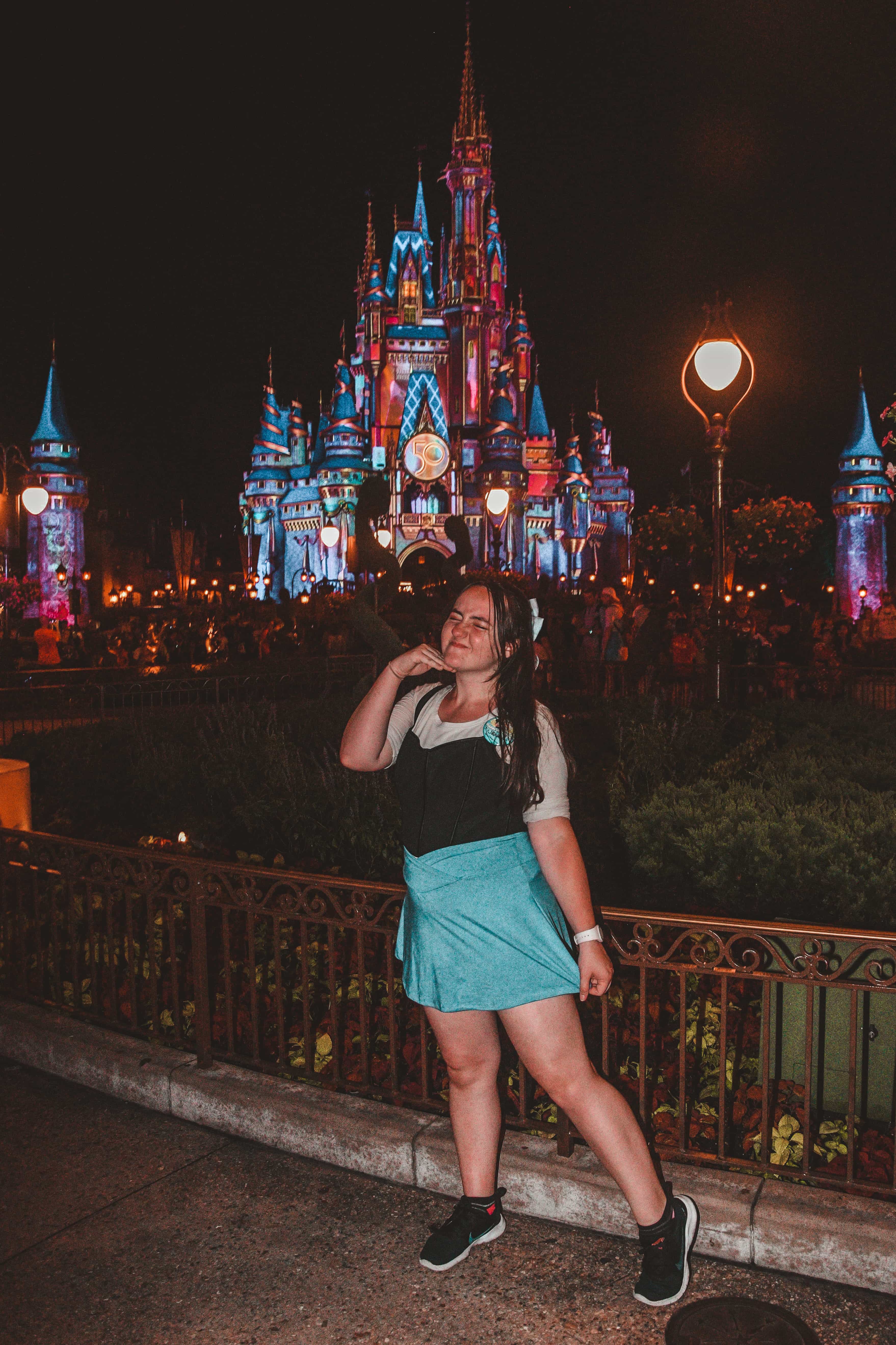 A girl posing in front of a castle at Walt Disney World while wearing a blue skirt, black shirt and black shoes. 