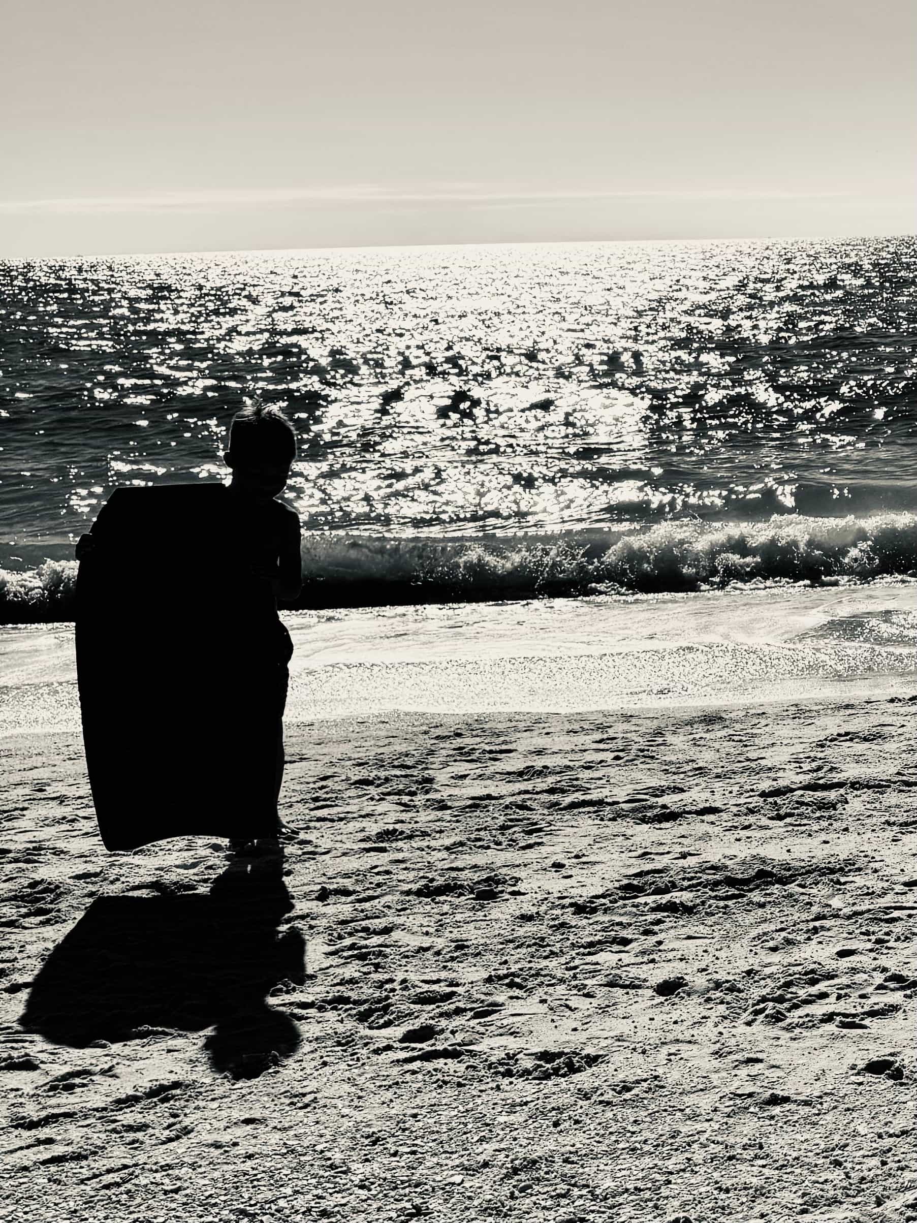 Black and white photo of a child holding a boogie board on the beach