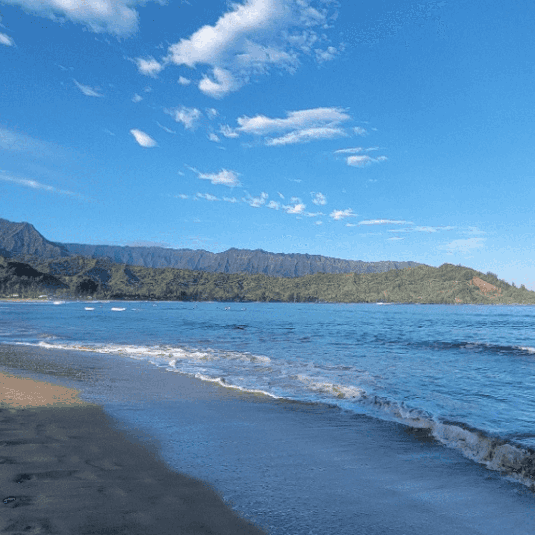 View of an empty beach and calm sea under sunny skies with green mountains in the distance