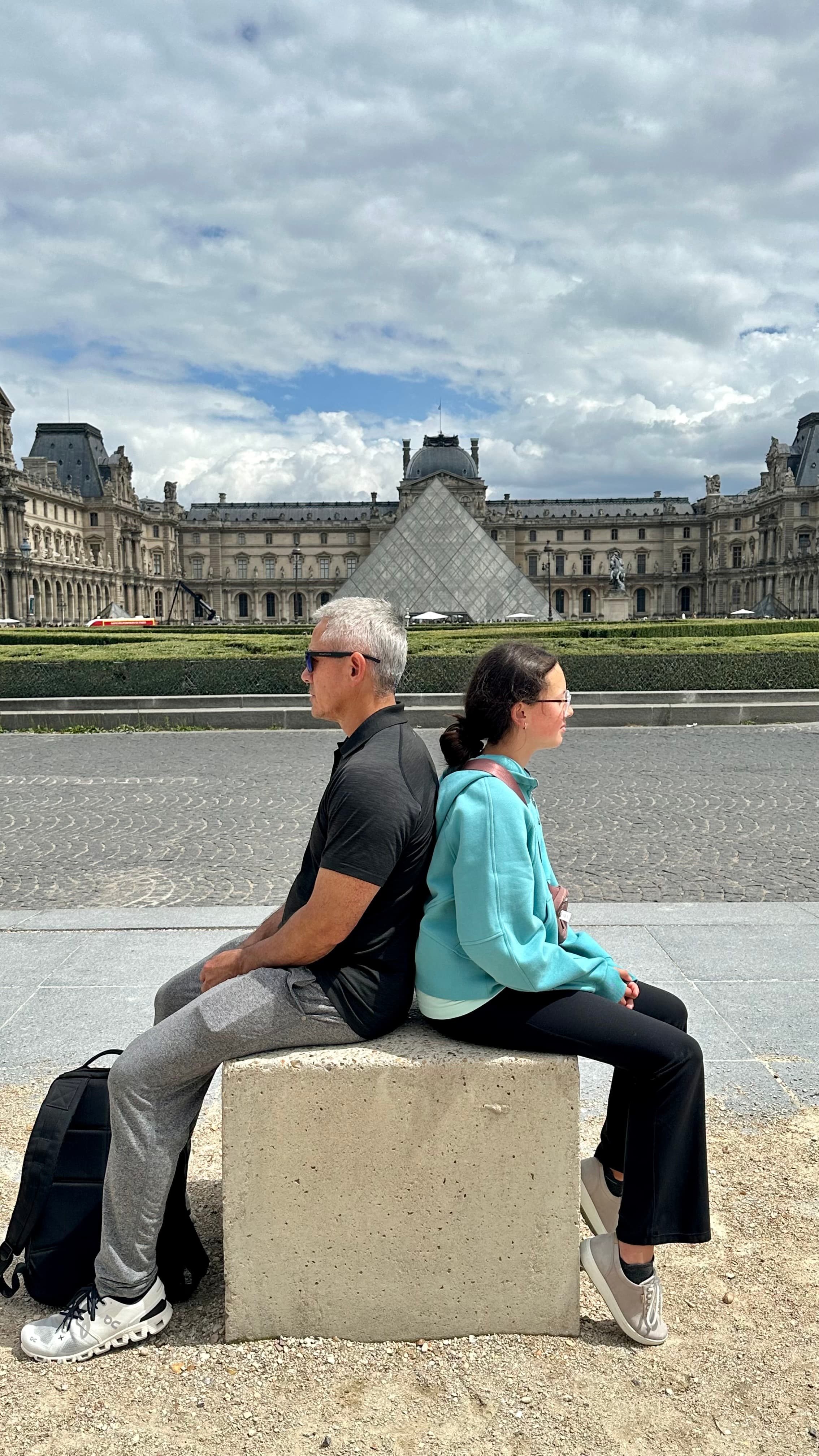 Two people sitting on a concrete structure with the Louvre Museum in the distance. 