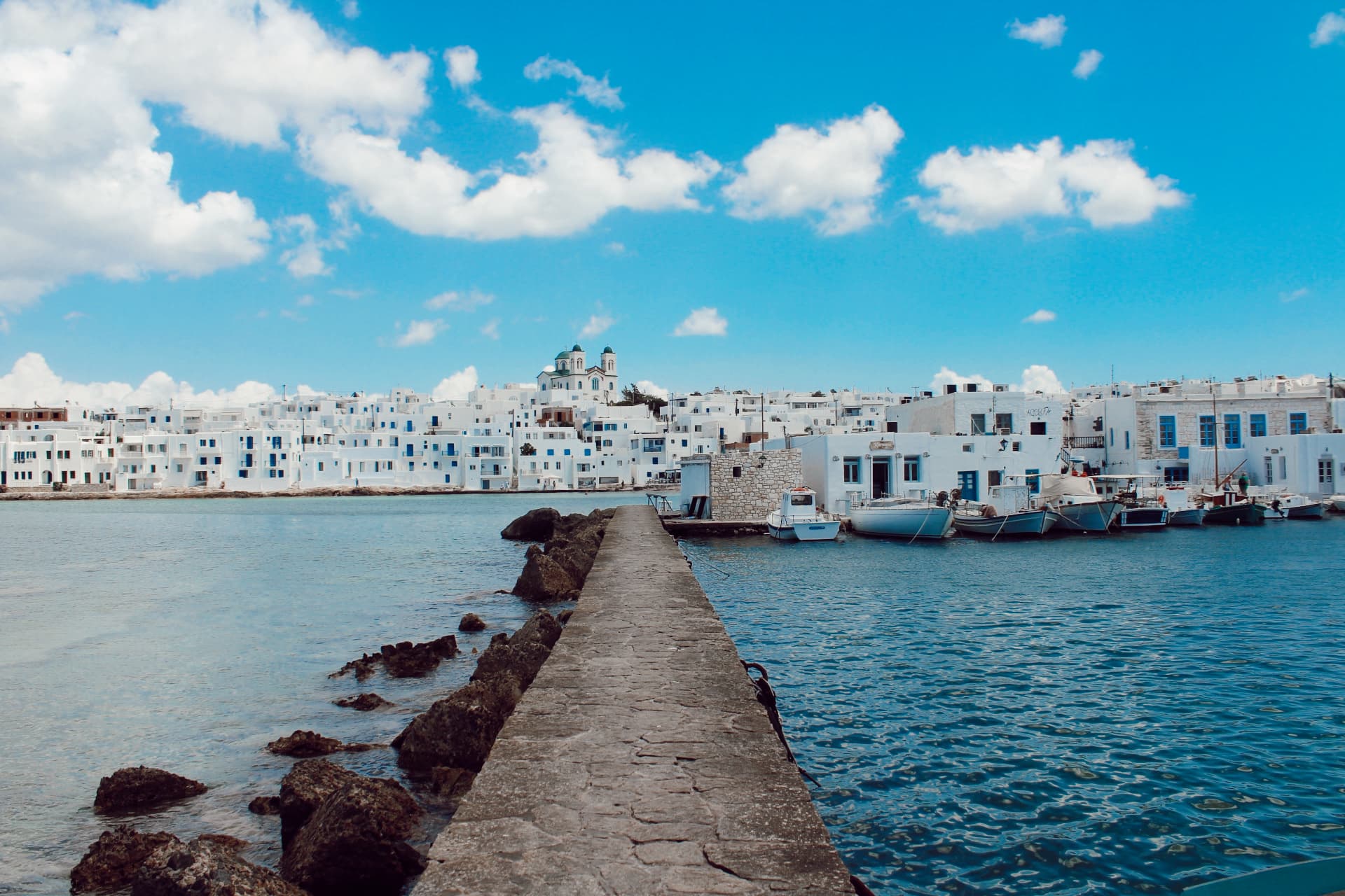 A view of a walking path next to the ocean with a town in the distance on a sunny day. 