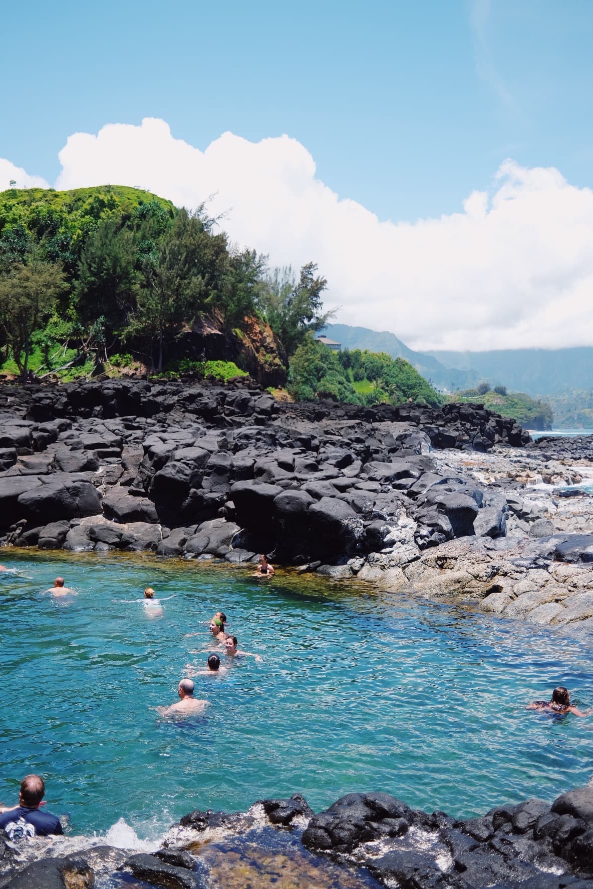 An image of a rocky beach with a swimming cove and foliage in the distance on a sunny day. 