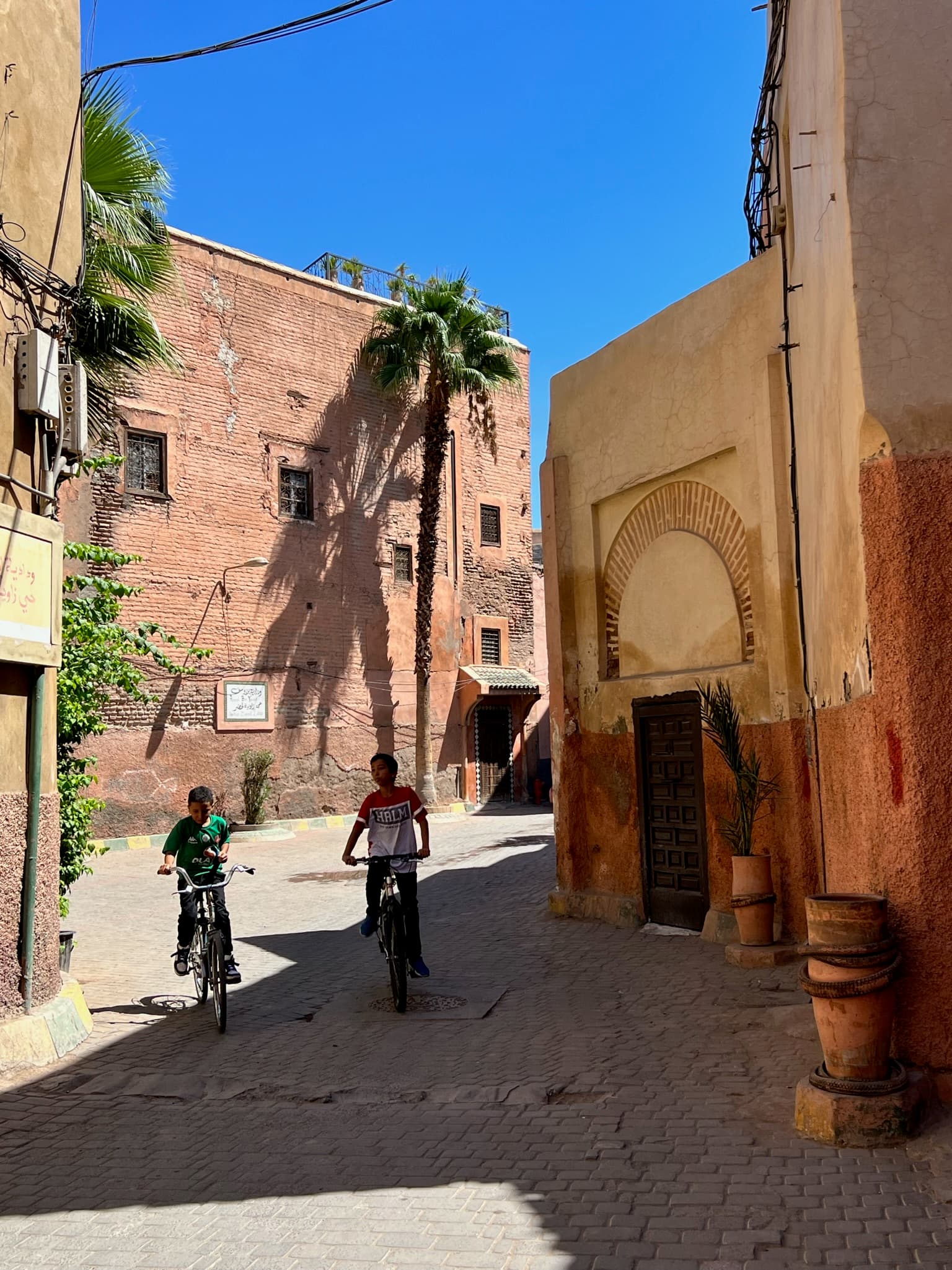 An image of a dirt road with people on bicycles with neutral colored buildings, foliage and flower pots during the day time.