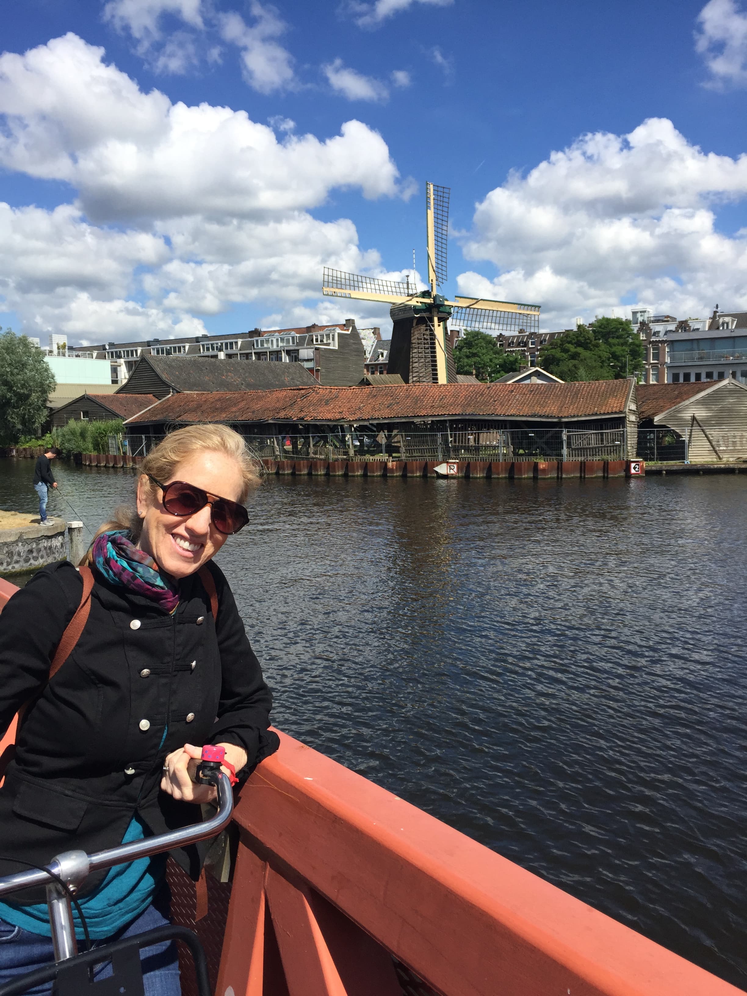 Jill on a bike on a bridge over a canal in Amsterdam, a windmill visible behind her