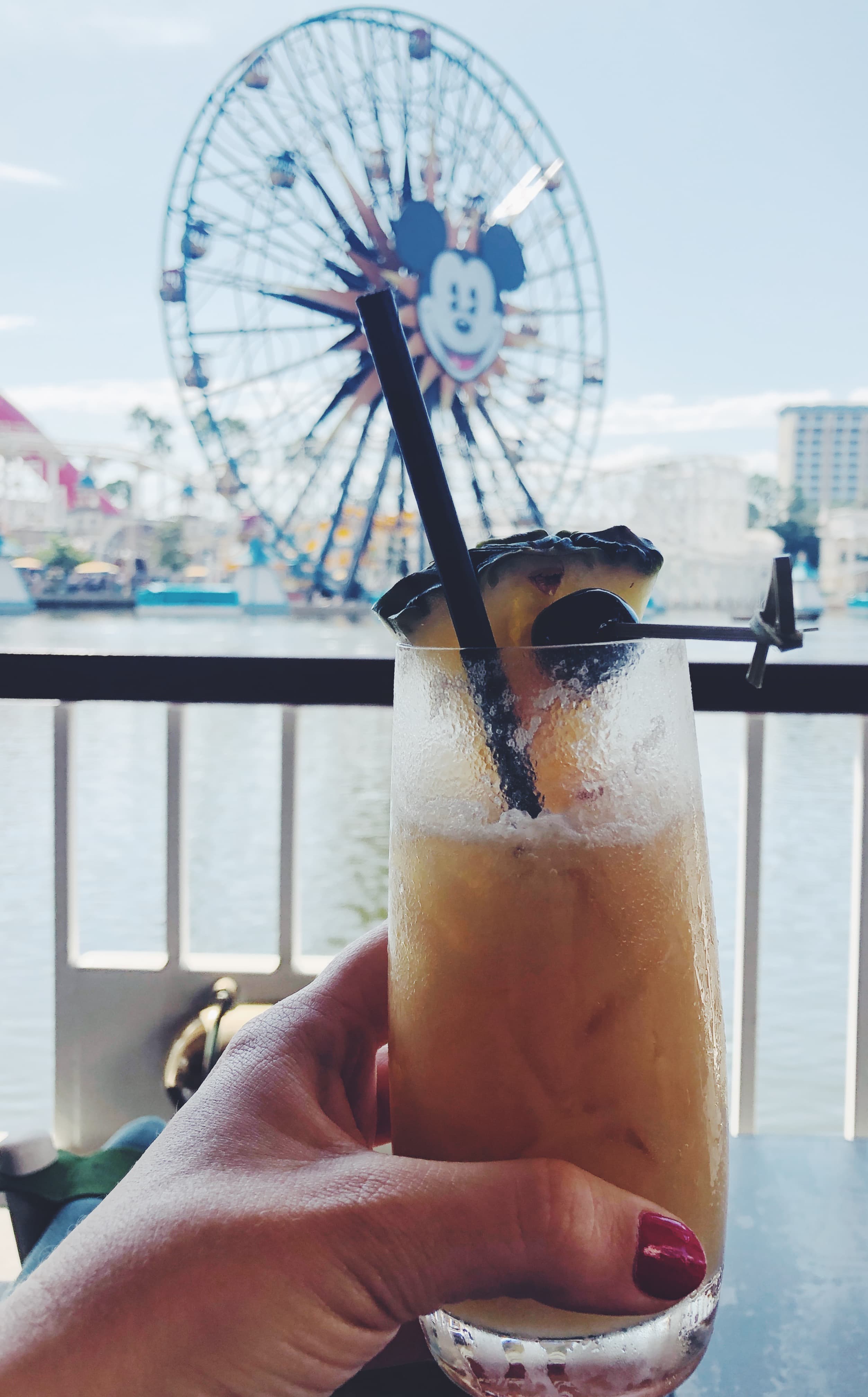 View of a hand holding a tropical cocktail with a slice of pineapple with a Mickey Mouse Ferris wheel visible behind it