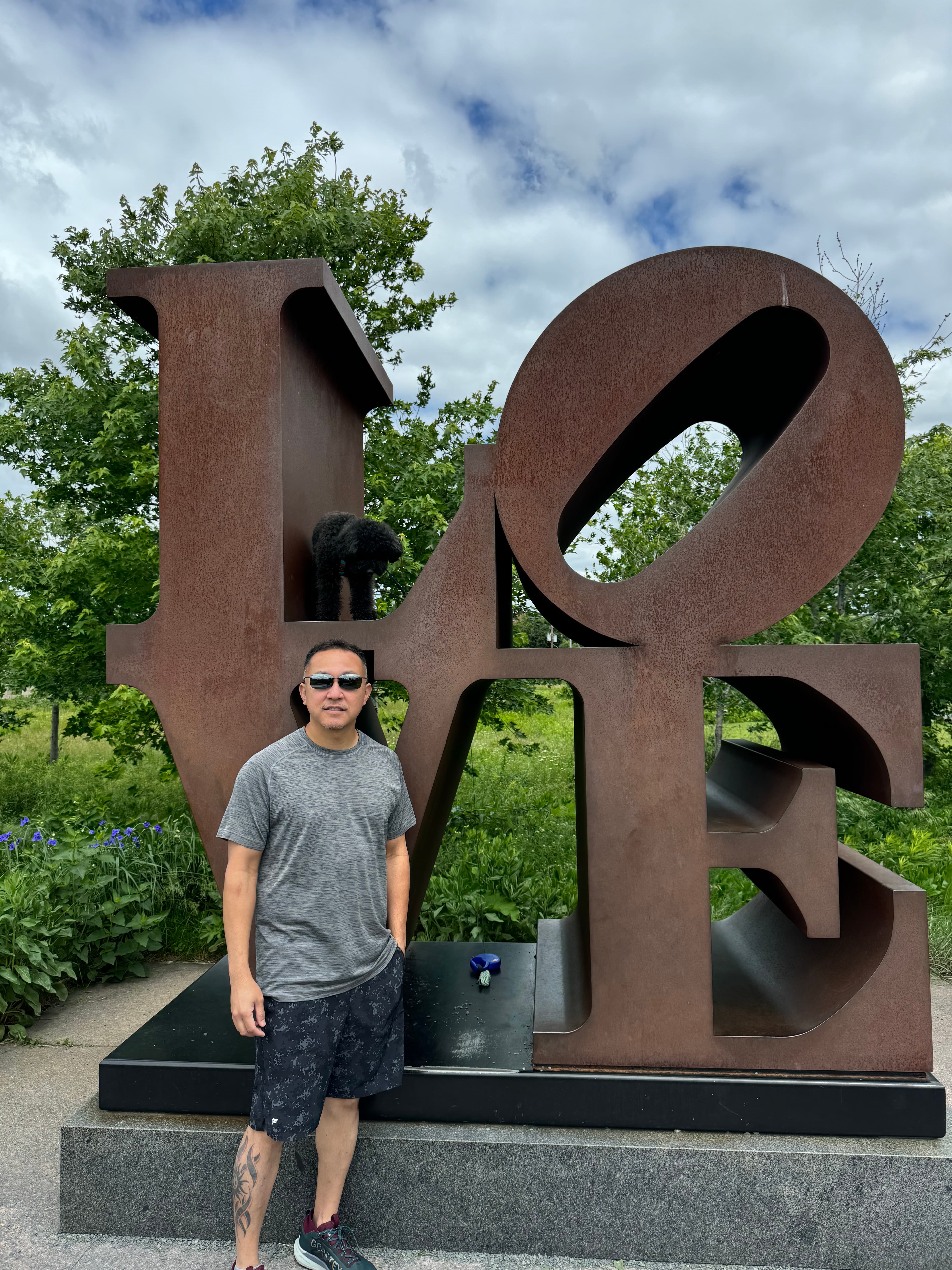 Scott in a grey t-shirt standing in front of a metal sign spelling “LOVE” on a cloudy day