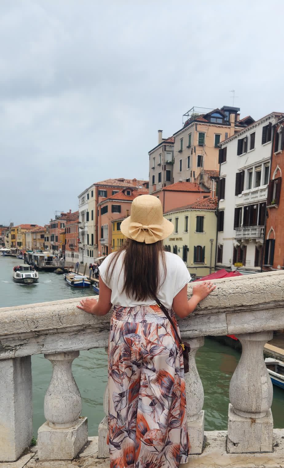 Chelsey wearing a hat while on a bridge over a canal lined with colorful buildings under cloudy skies
