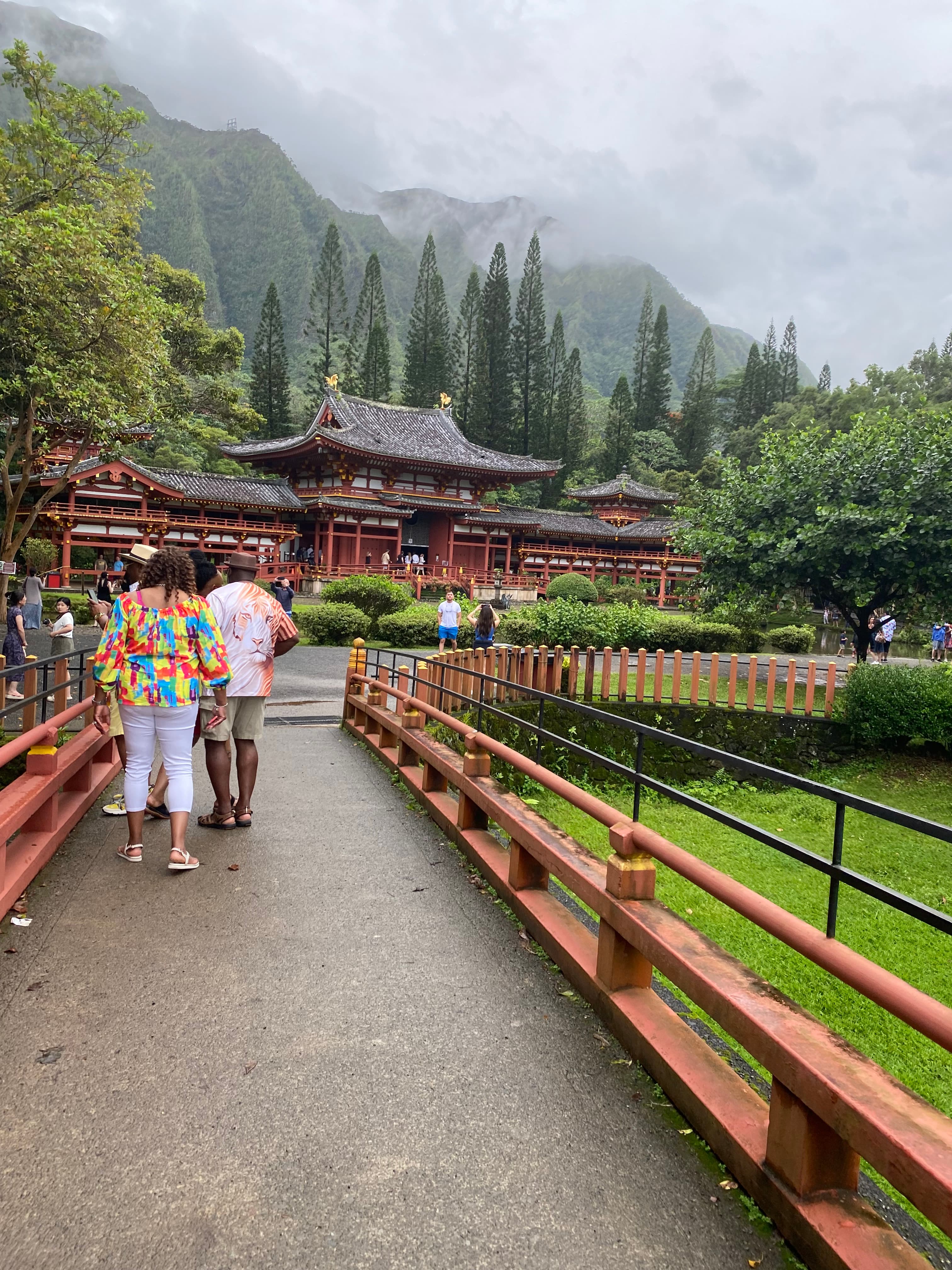 People on a pathway leading towards a large temple with trees and mountains visible behind it on a cloudy day