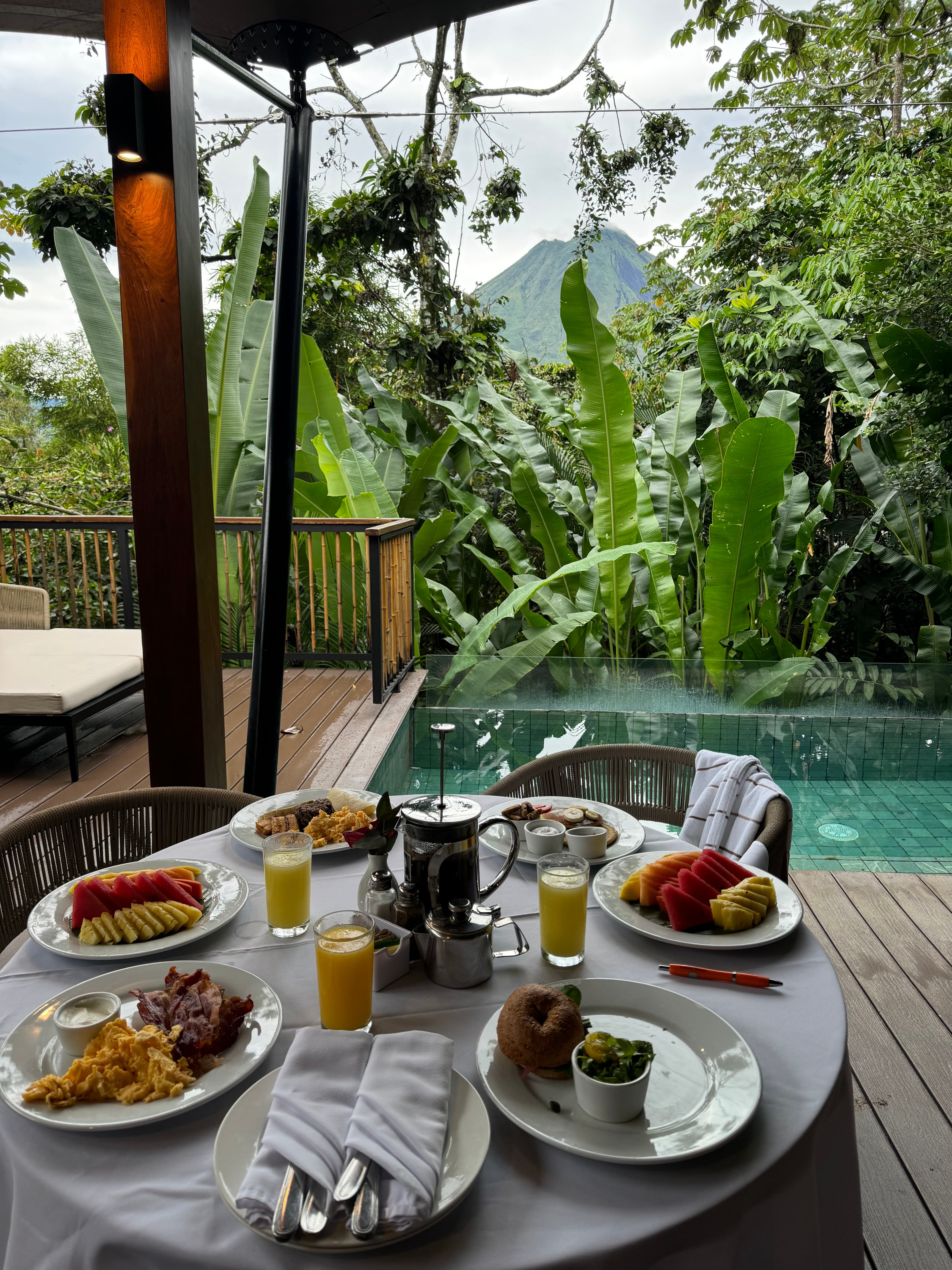 View of a beautifully set breakfast table with several plates outdoors overlooking a pool and tropical plants