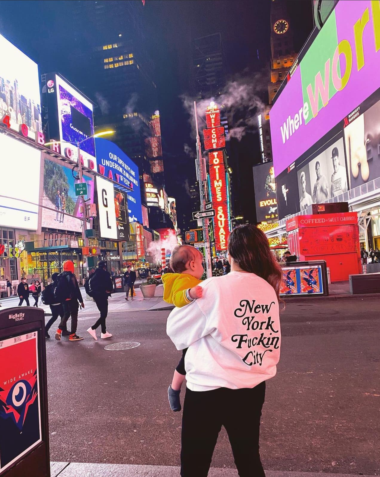 Advisor holding a baby in a white sweatshirt walking through Times Square in NYC at night