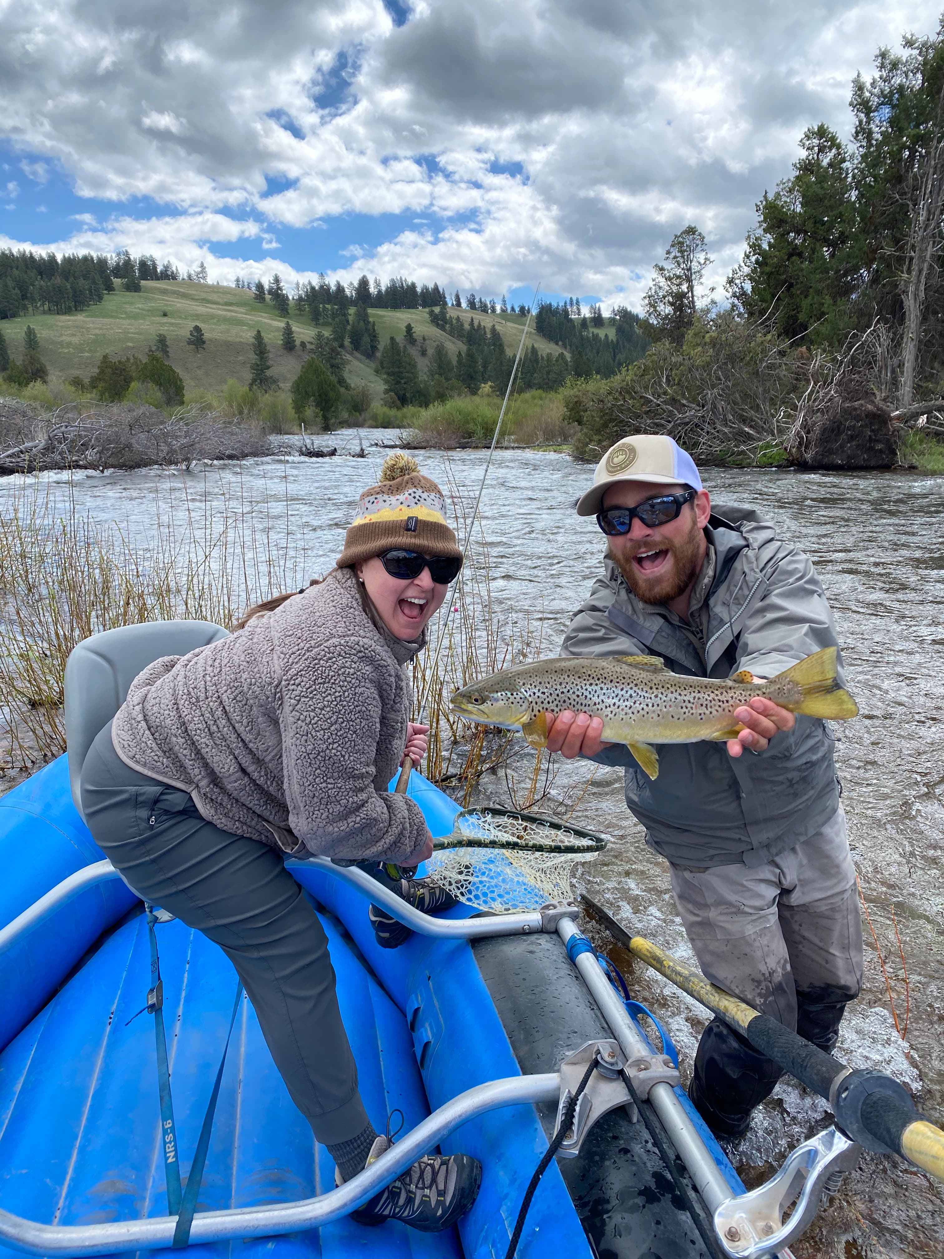 Tanya in a blue boat on a riverbank with a man holding a fish next to her