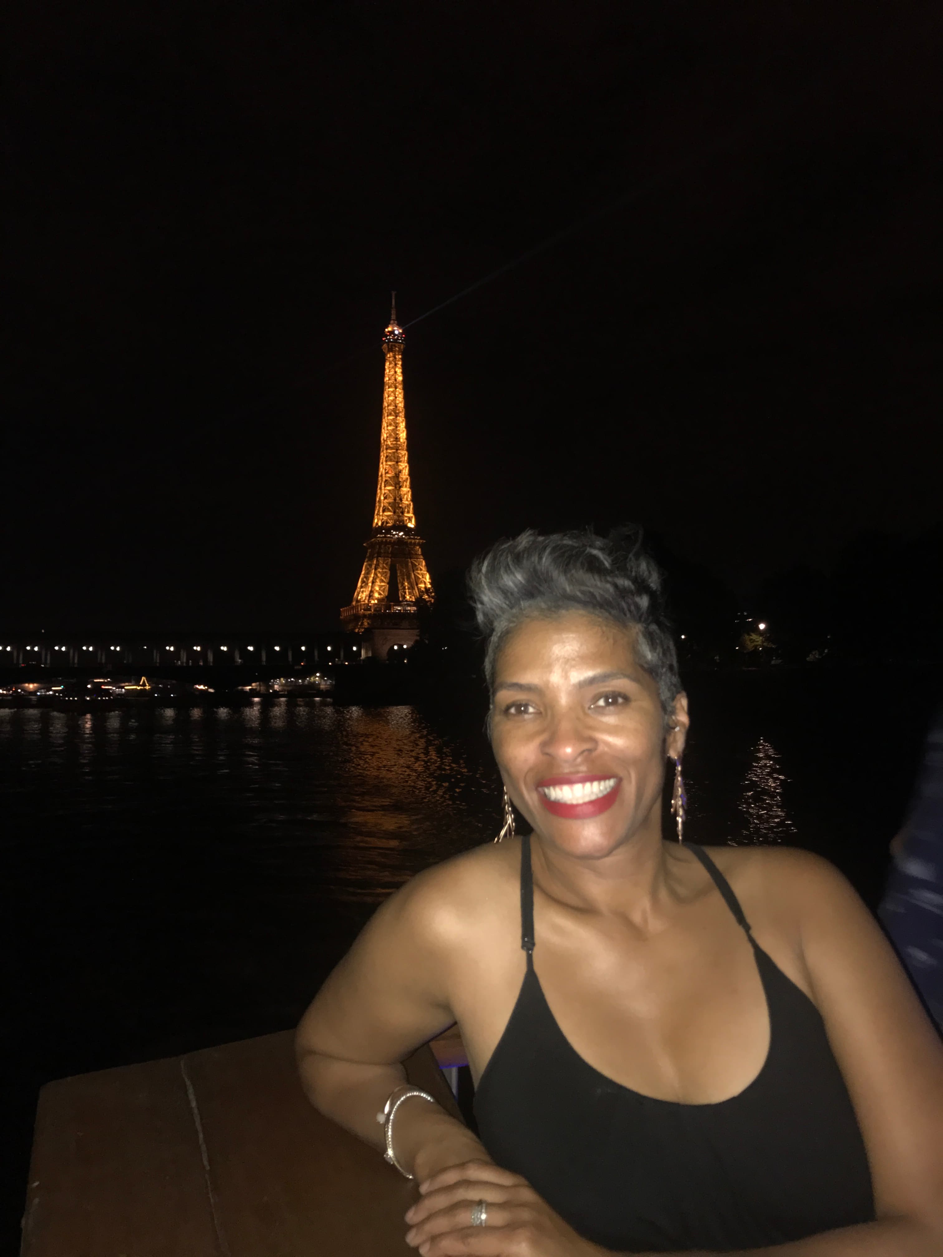 Katrina in a black dress smiling for a photo with the Eiffel Tower lit up in the distance behind her at night