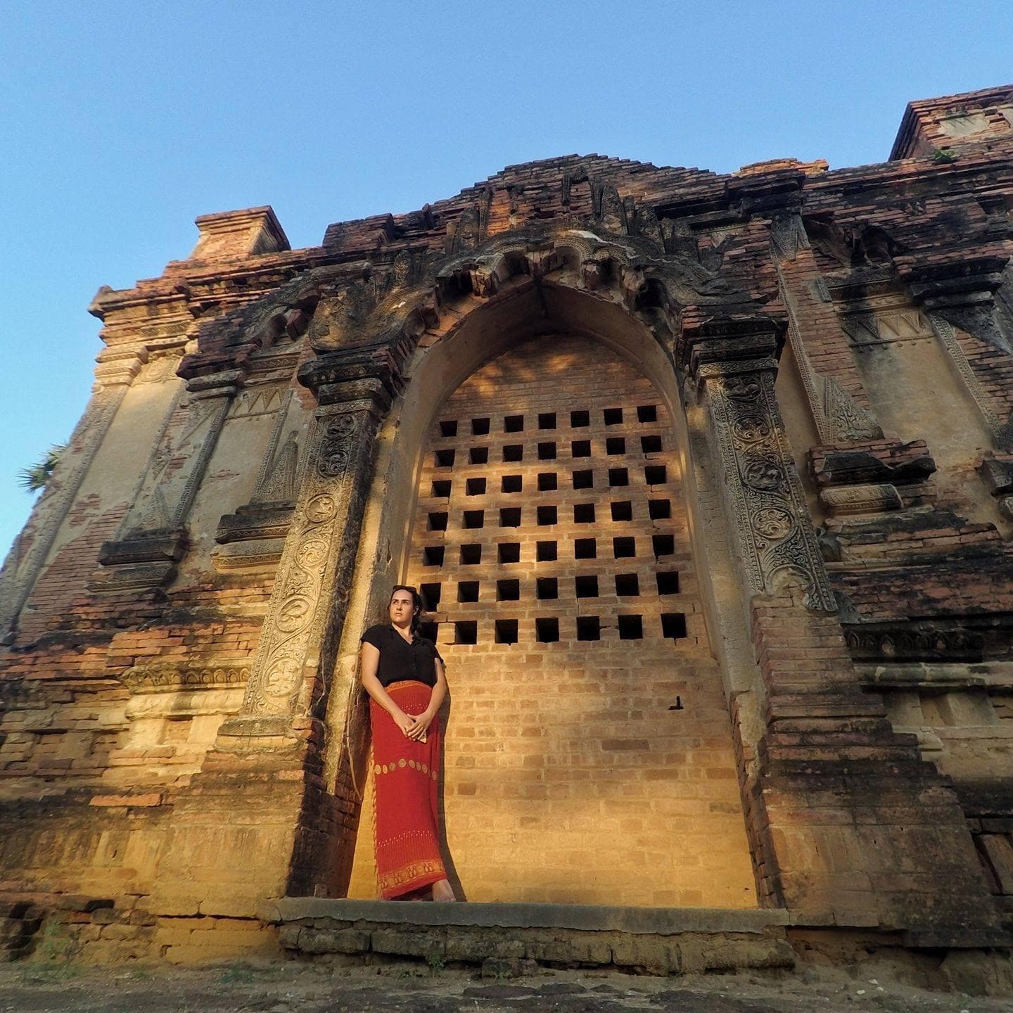 View of advisor in a long red skirt leaning against the doorway to an ancient building under clear skies