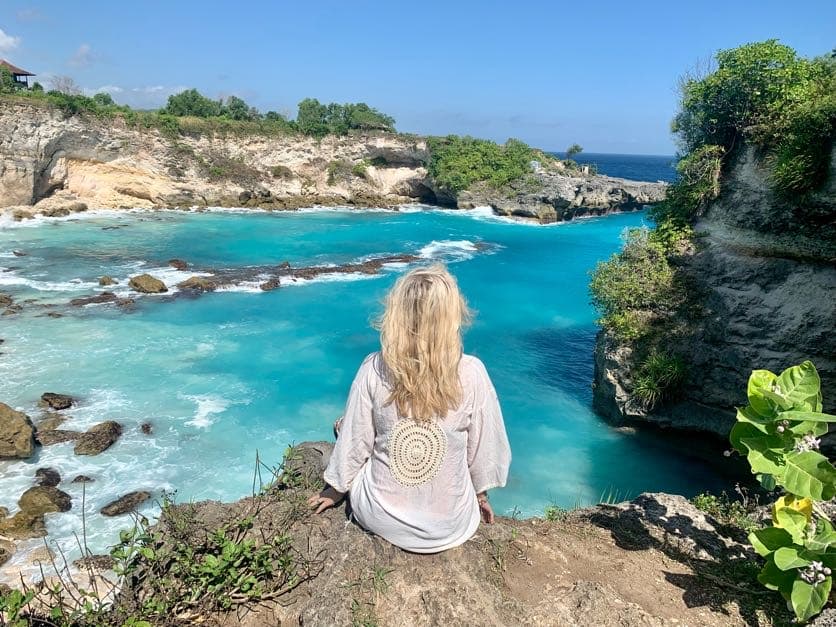 Advisor sitting on a rock ledge looking out over a teal blue ocean cove with cliffs and plants surrounding it on a sunny day.