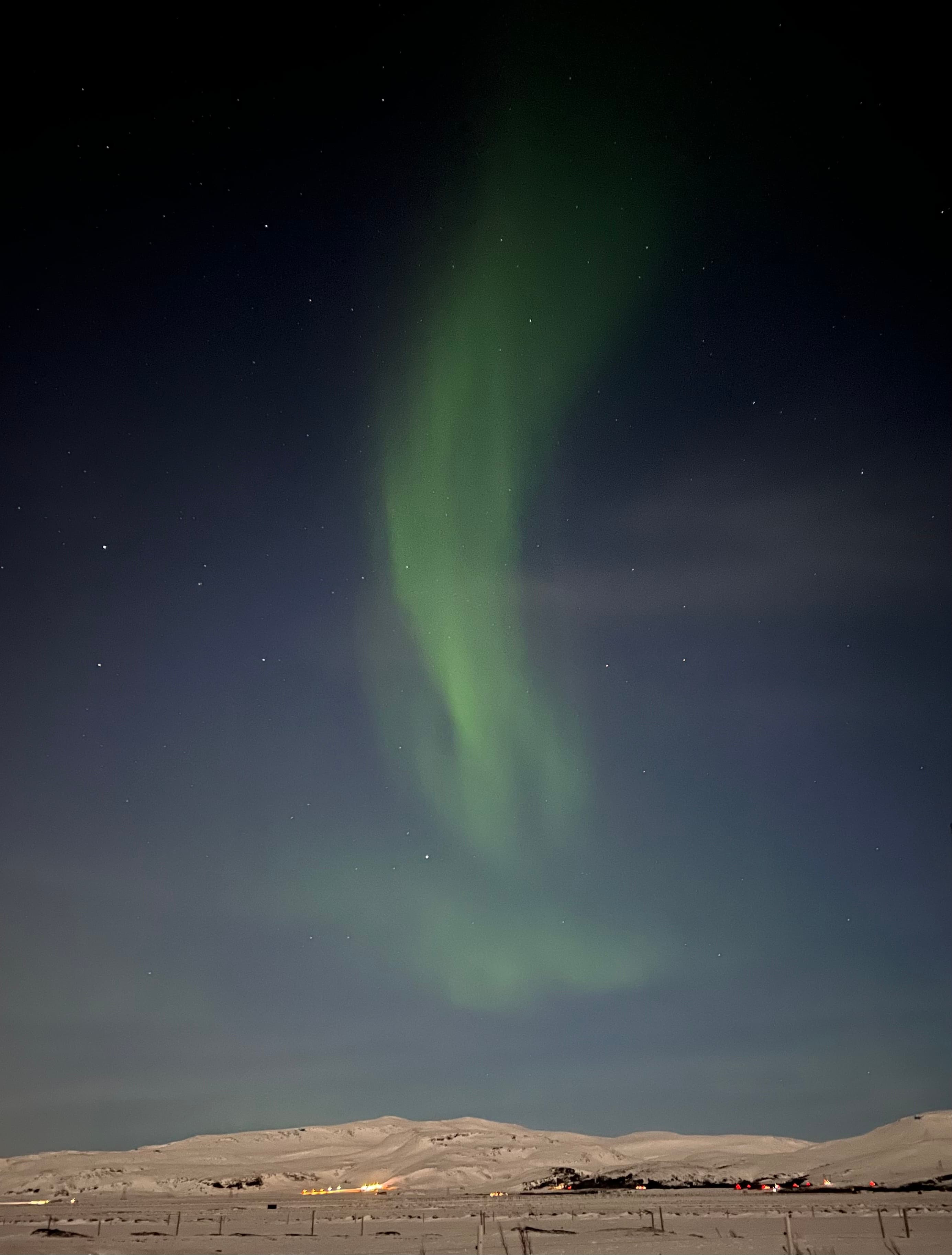 Northern lights dance in the sky as a green hue reflects off clouds over snow-capped mountains. 
