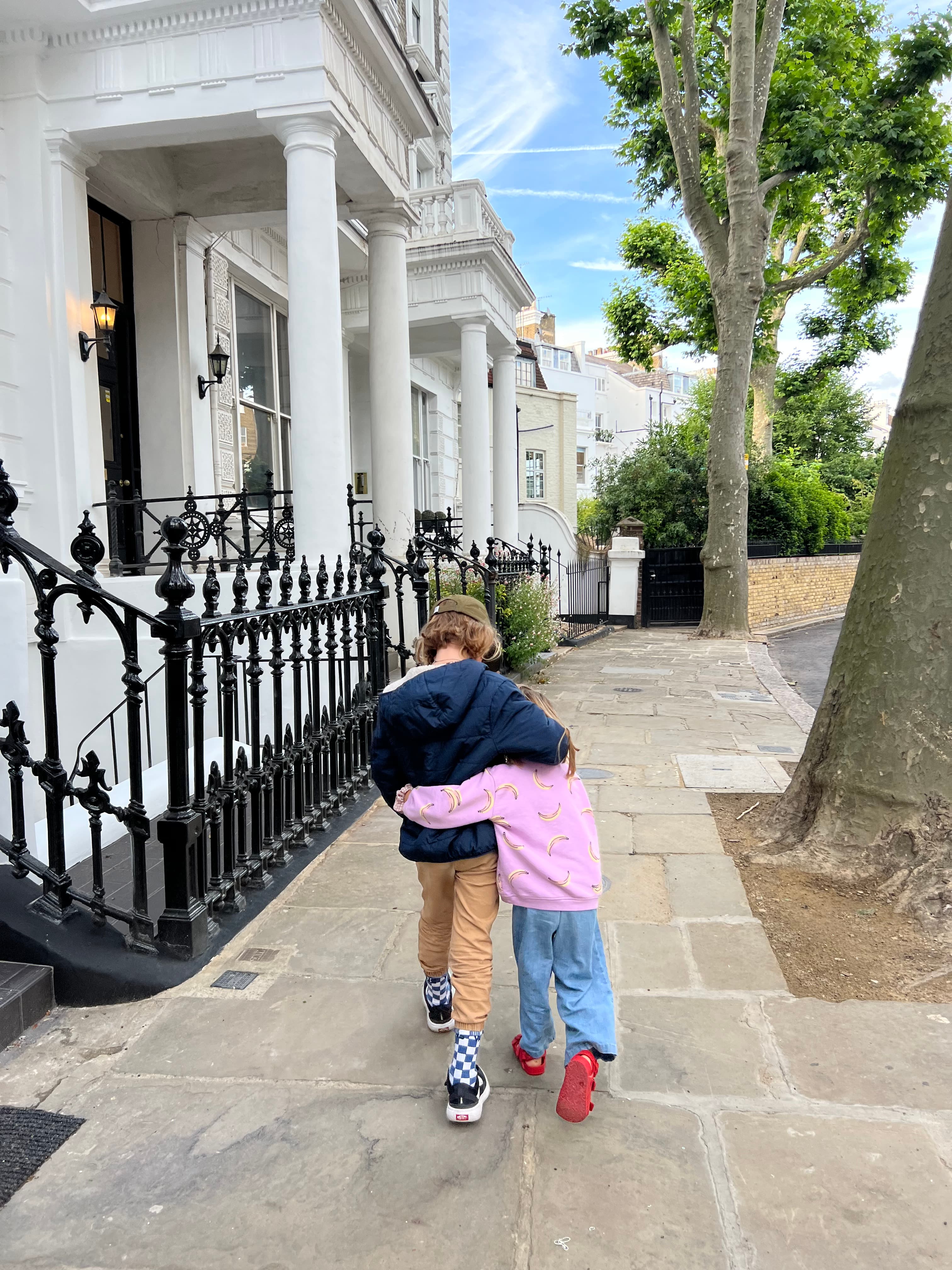 Two children walk down a paved street with their arms around one another.