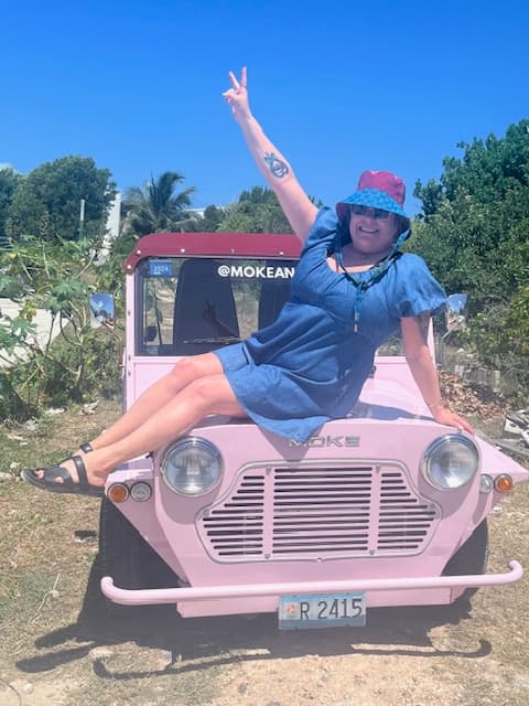 A woman wearing a pink dress is perched on a pink car outdoors during the daytime.