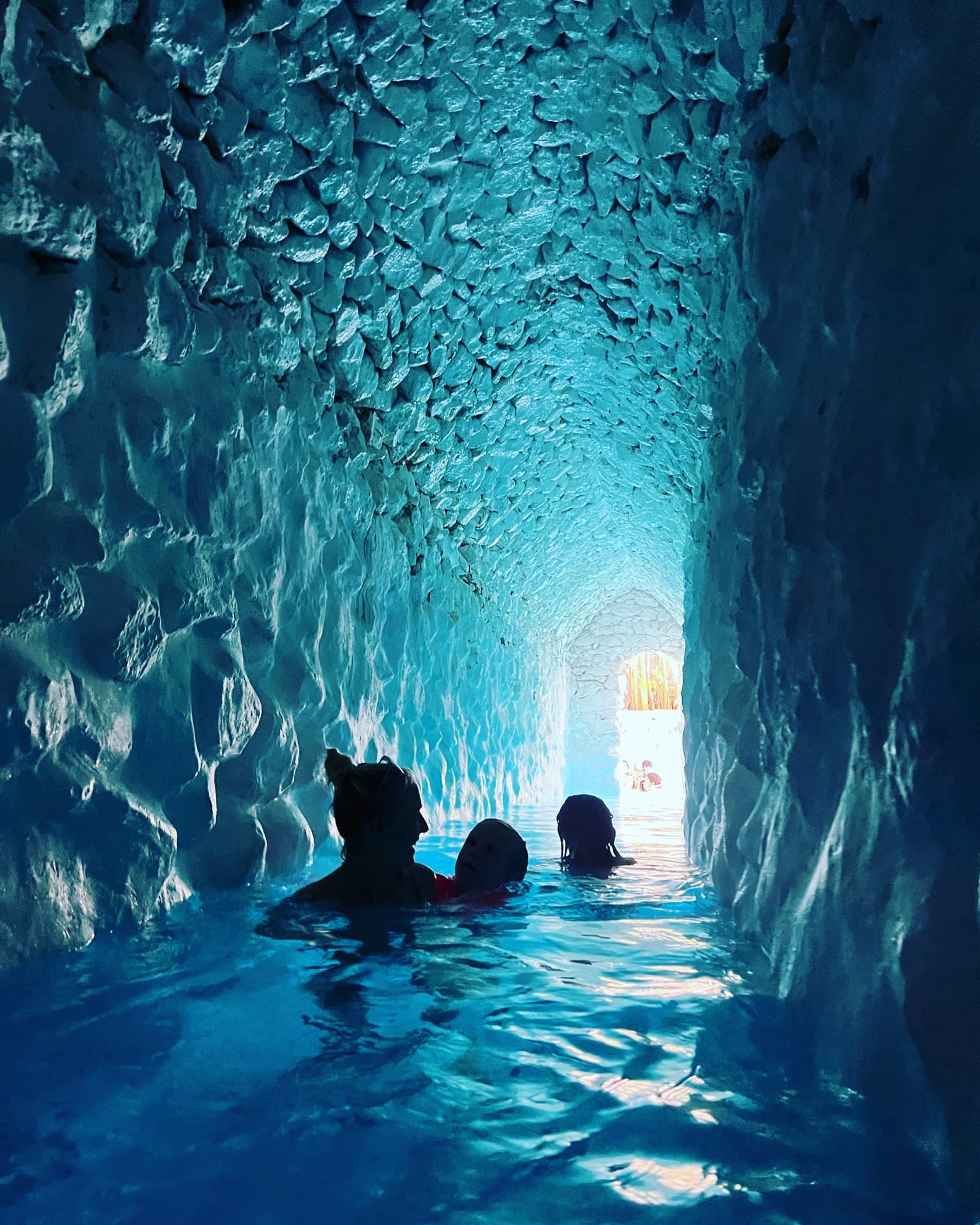 View of three people in a narrow pool in a cave with stone ceilings