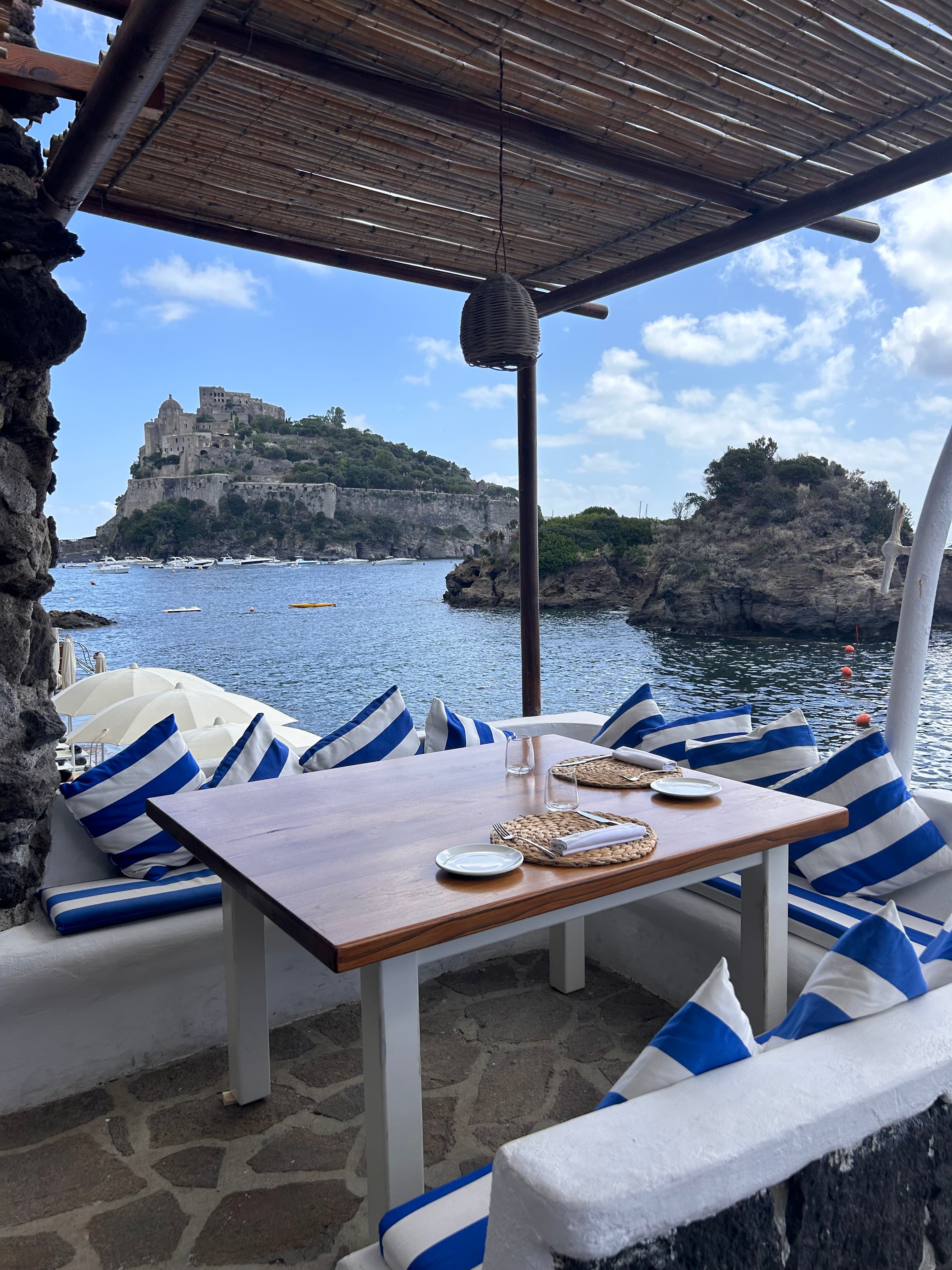 View of a wooden table set with plates and white and blue striped pillows by the sea