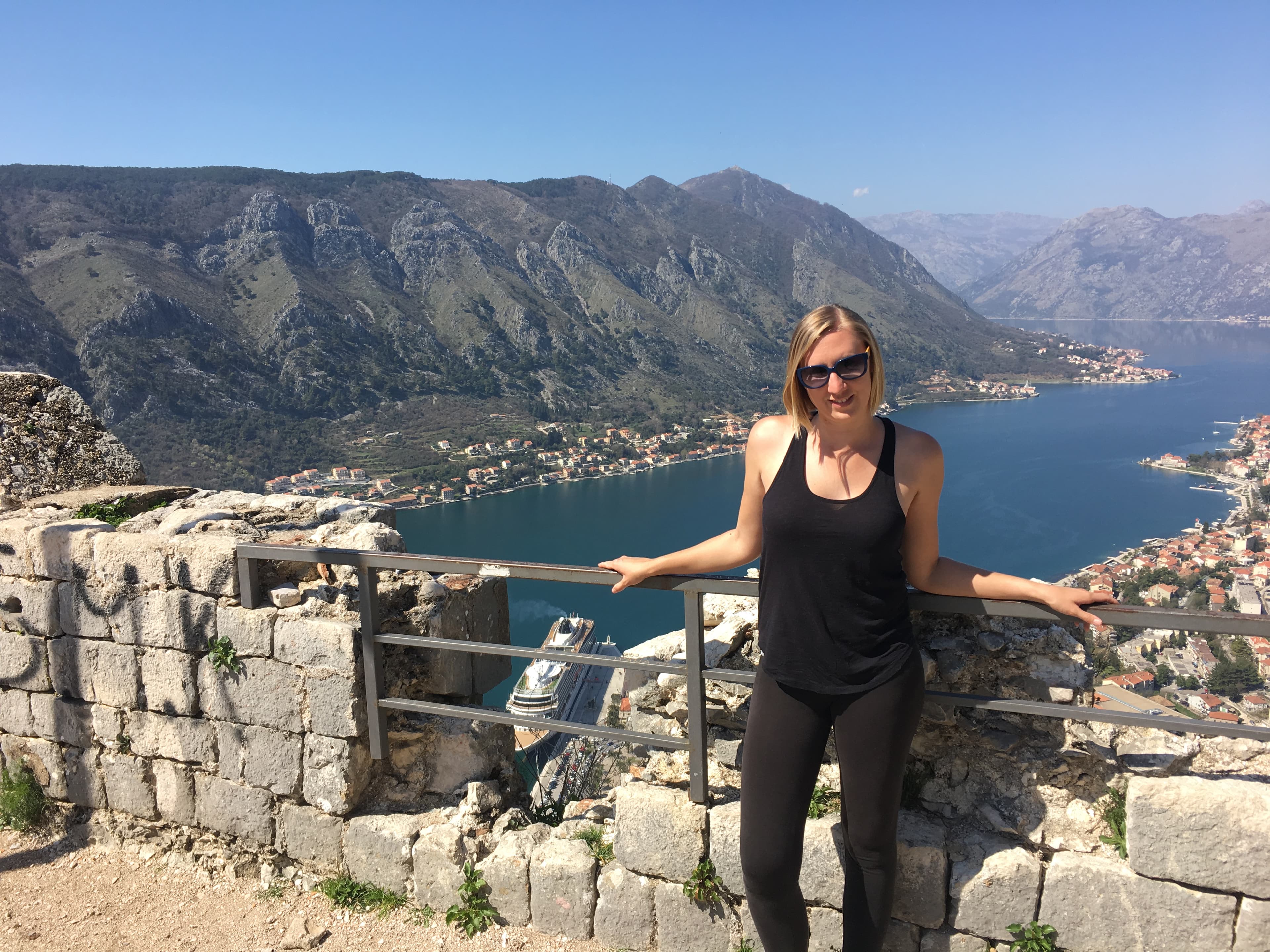 Cassie posing in a black tank top against a railing with mountains and water in the background during the daytime.