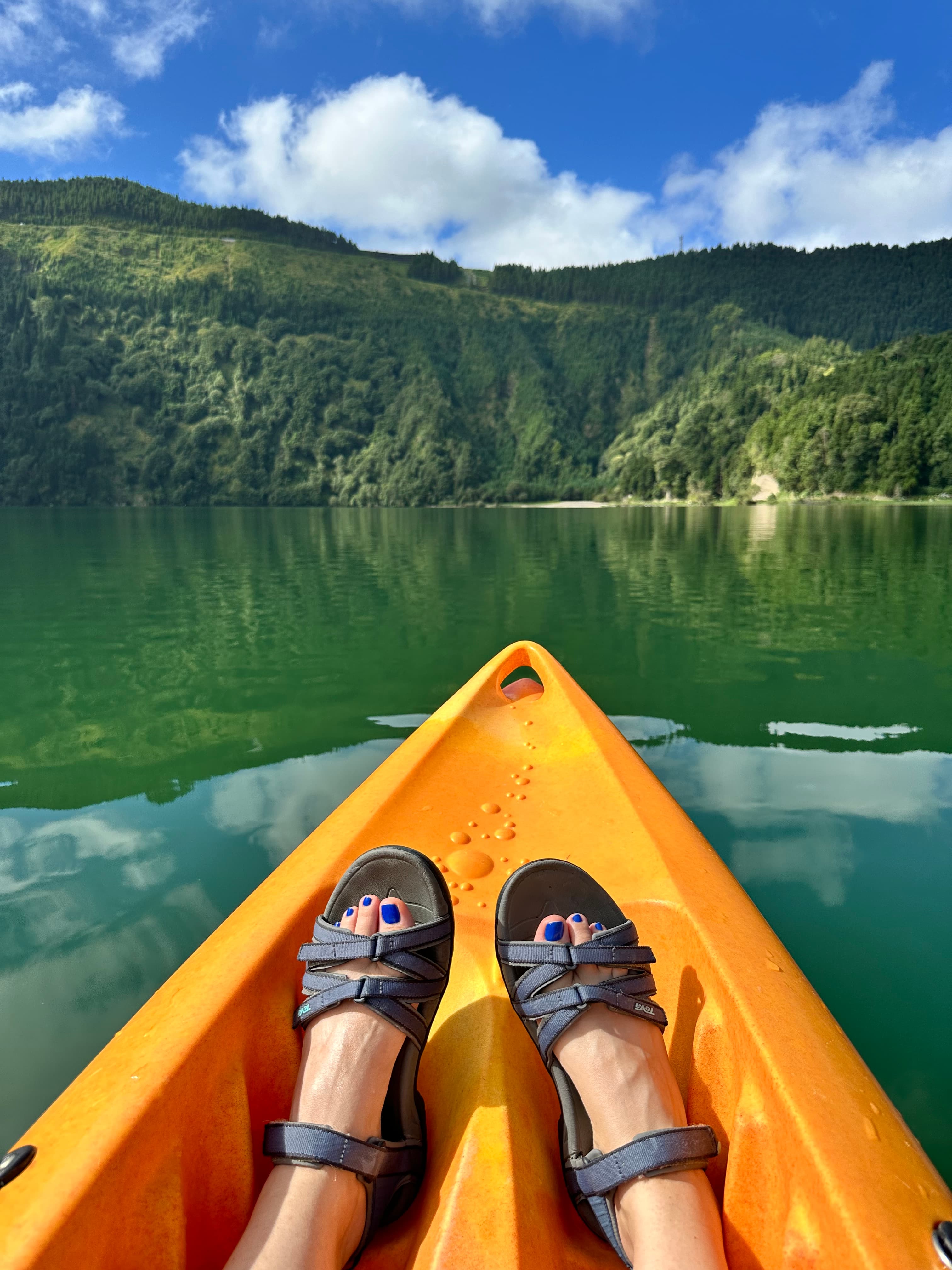 View of advisor’s feet in sandals at the front of a yellow kayak on a glassy green lake surrounded by green hills