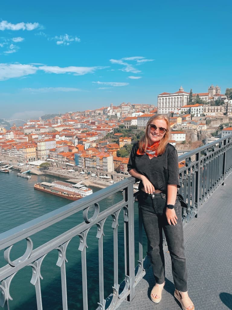 Picture of Merritt wearing a black top and black jeans leaning on a bridge in front of a city view and blue water under the cloudy blue sky