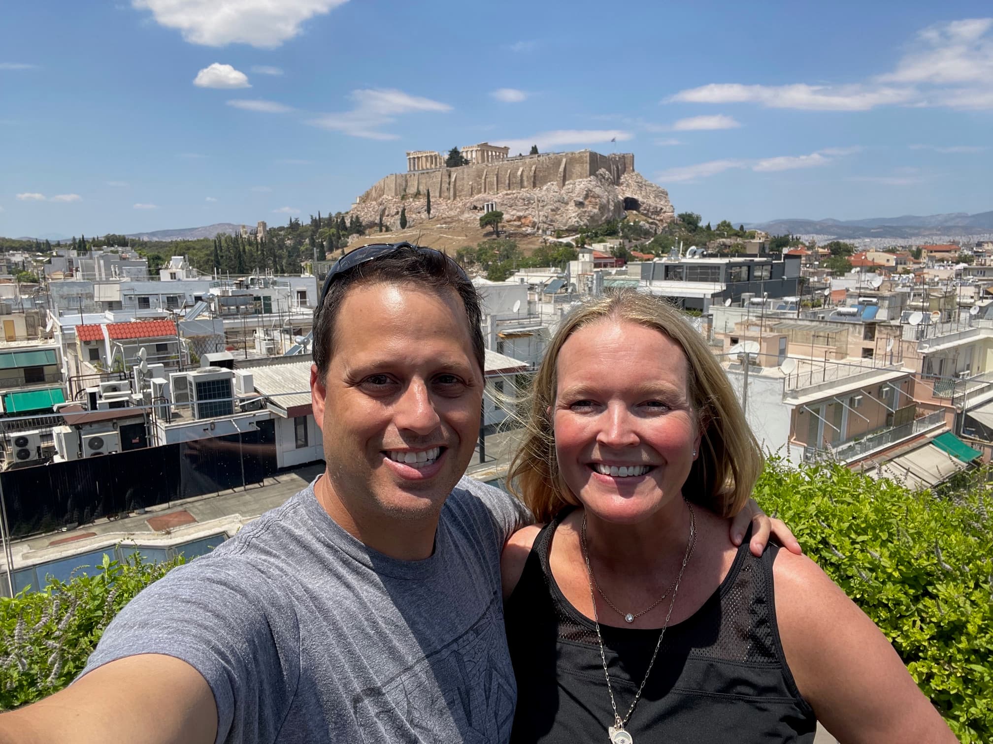 A couple posing for a selfie with a city and Greek temple in the background