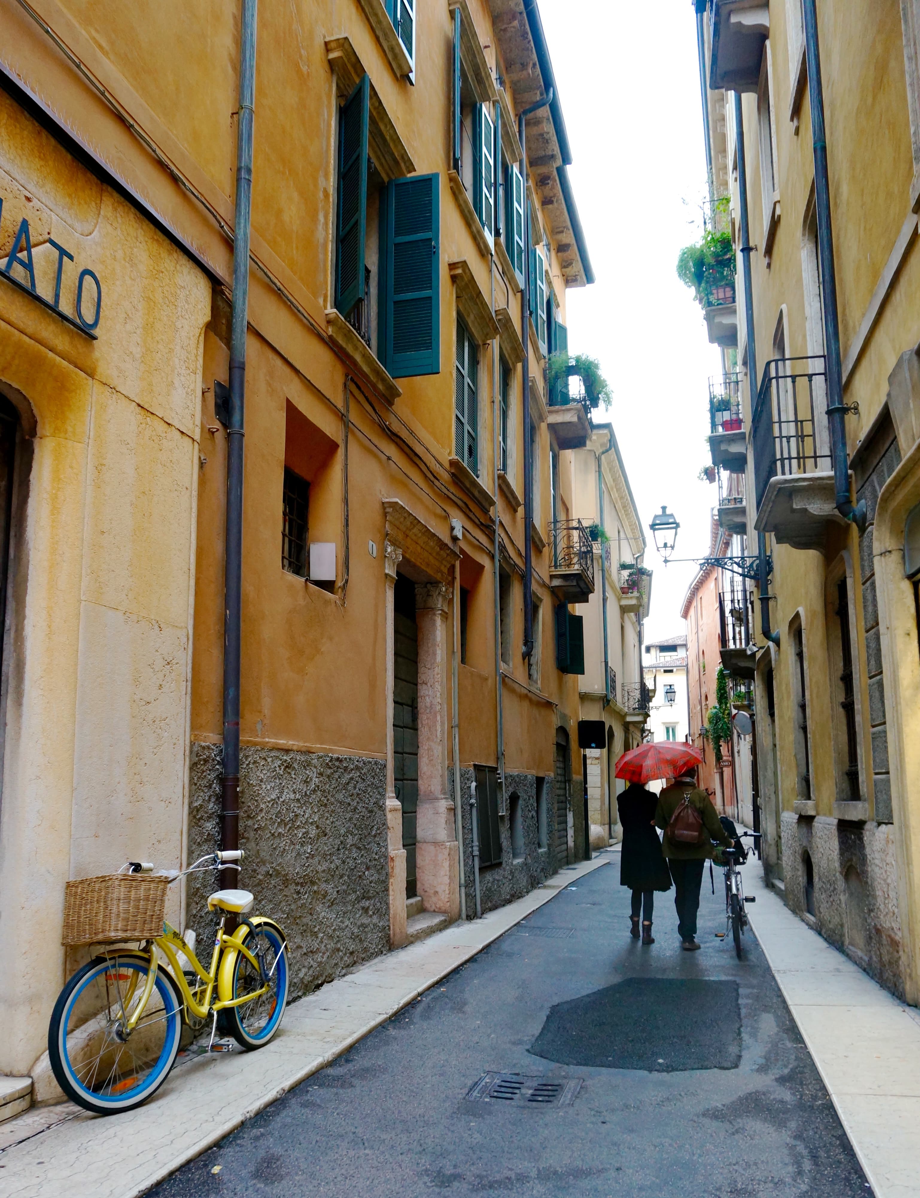 Two people walking down a European city street with a red umbrella in hand, yellow buildings with blue shutters and a bicycle in the surrounding areas