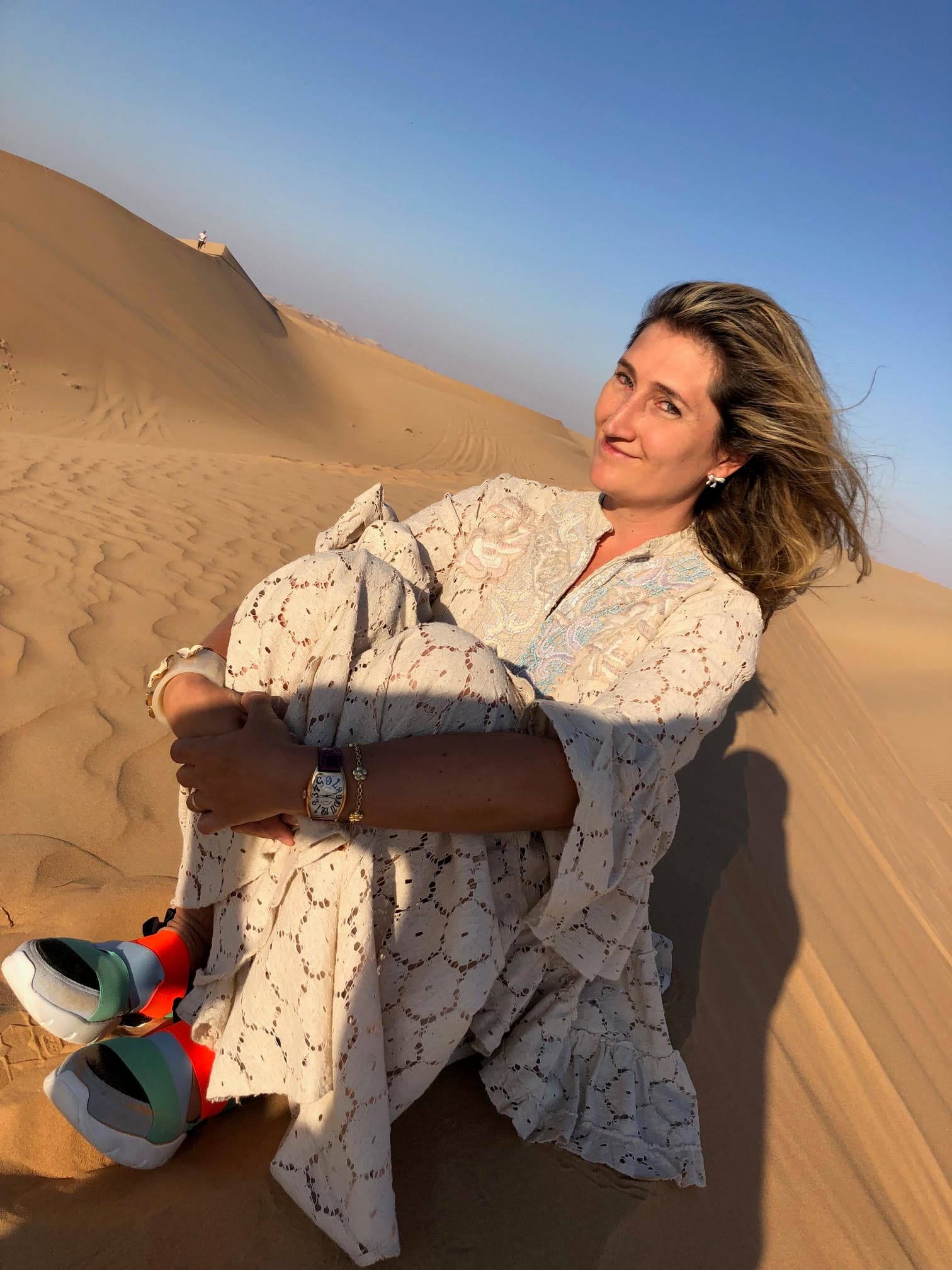 Favia sitting on the ground in the desert sand, hugging her knees, with her hair flowing in the breeze.