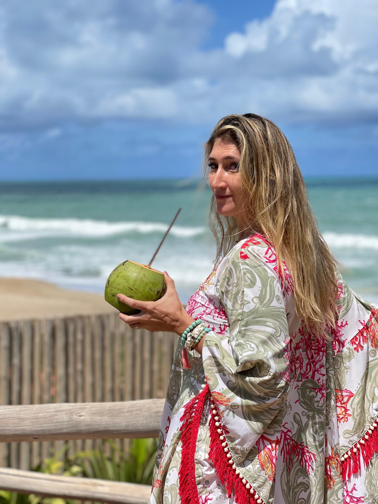 Favia on the beach draped in a flowing garment, holding a coconut, and about to take a sip from it.