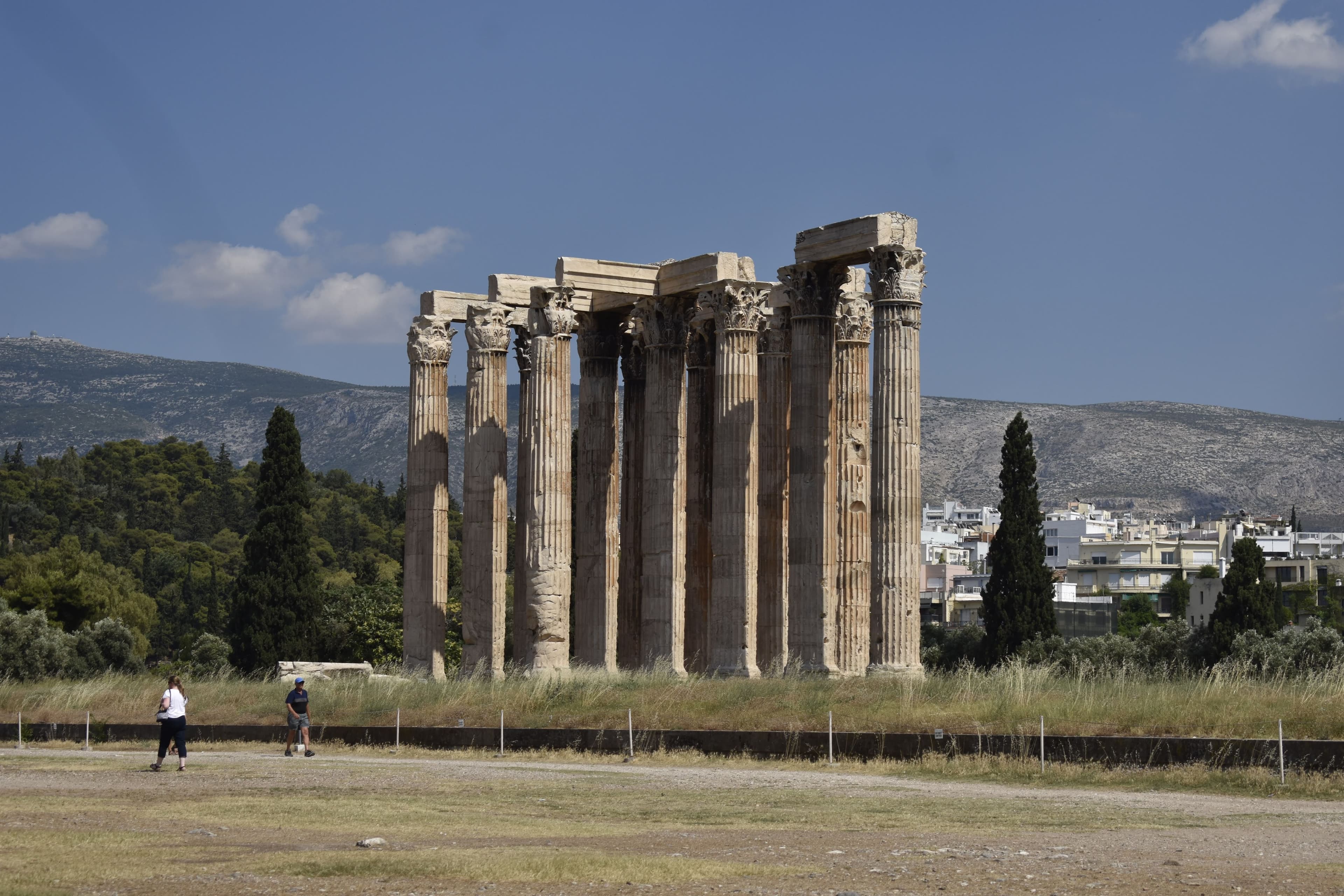 A photo of a Greek temple surrounded by trees and grass with two people standing in the front and a mountain in the background