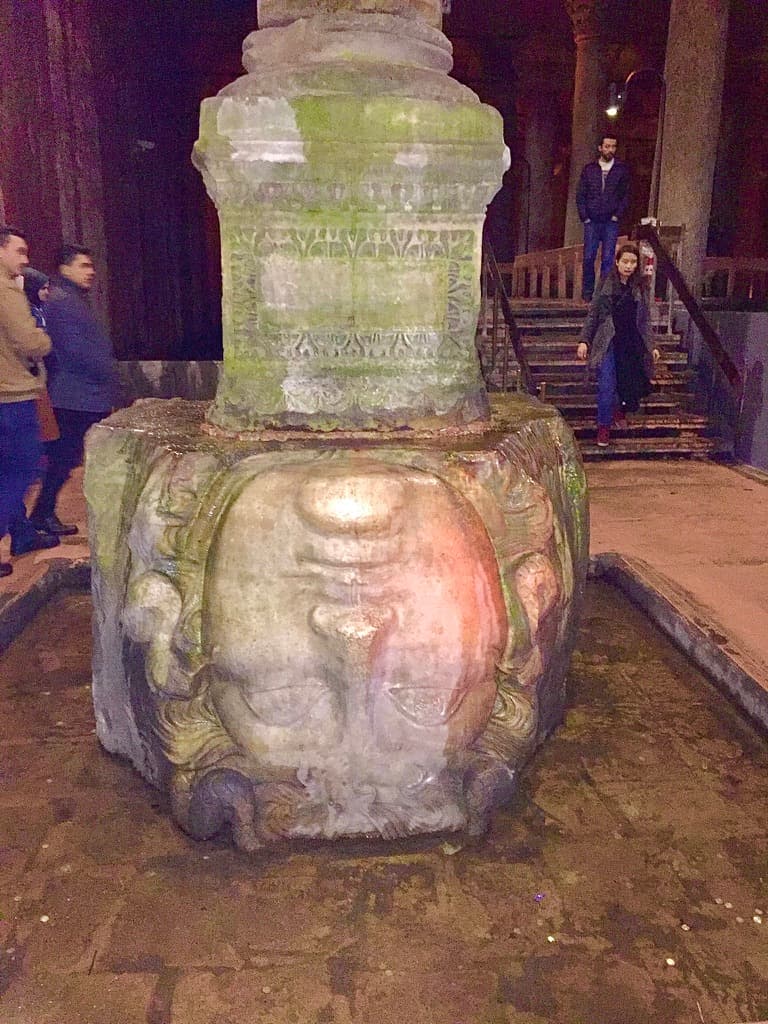 A picture of a Medusa head statue inside of a building with a staircase and people walking in the background