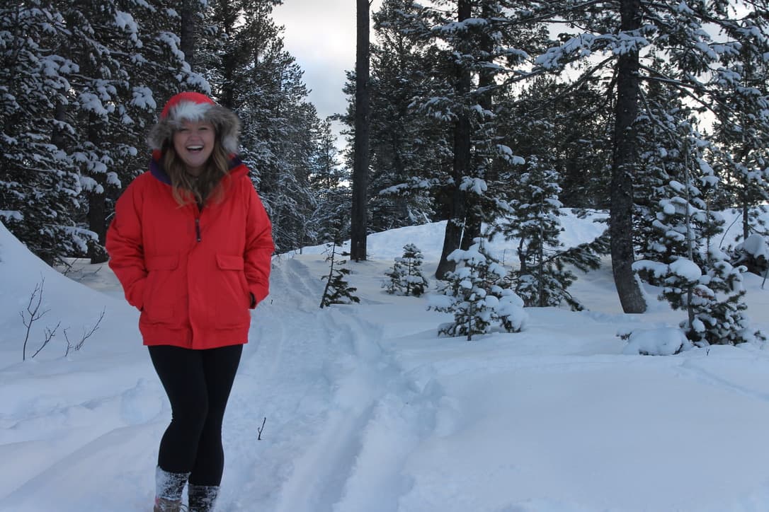 Picture of Merritt wearing a red jacket and smiling on a snowy trail surrounded by pine trees outside