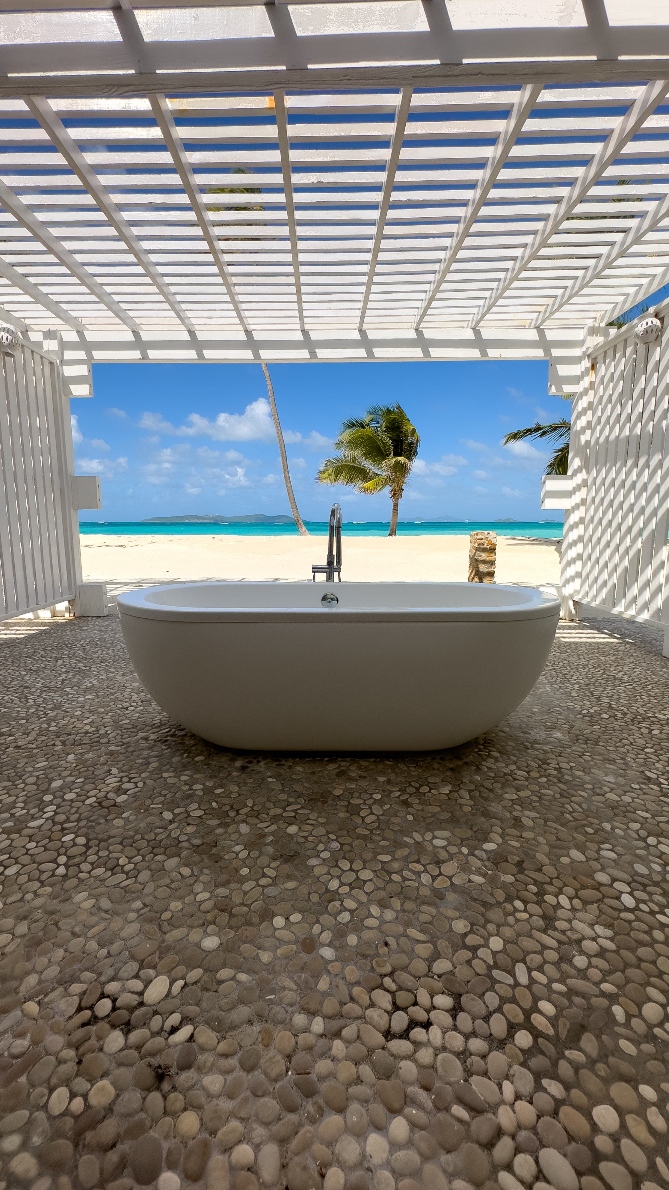 A white bath tub on a stone floor with a beach, palm tree and ocean view in the background