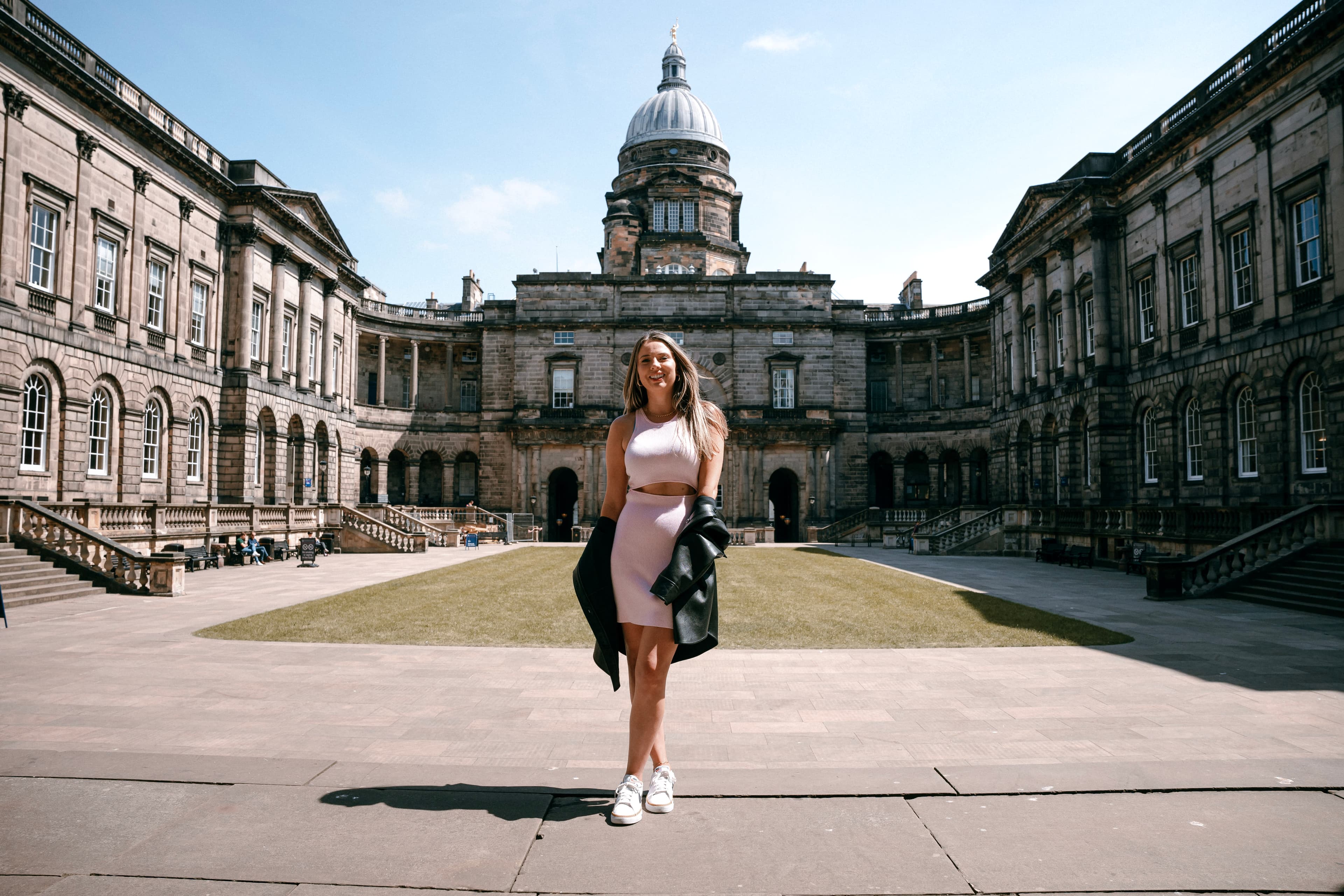 Emily in a skirt and tank top posing for a photo in the middle of a courtyard with The University of Edinburgh buildings surrounding it.