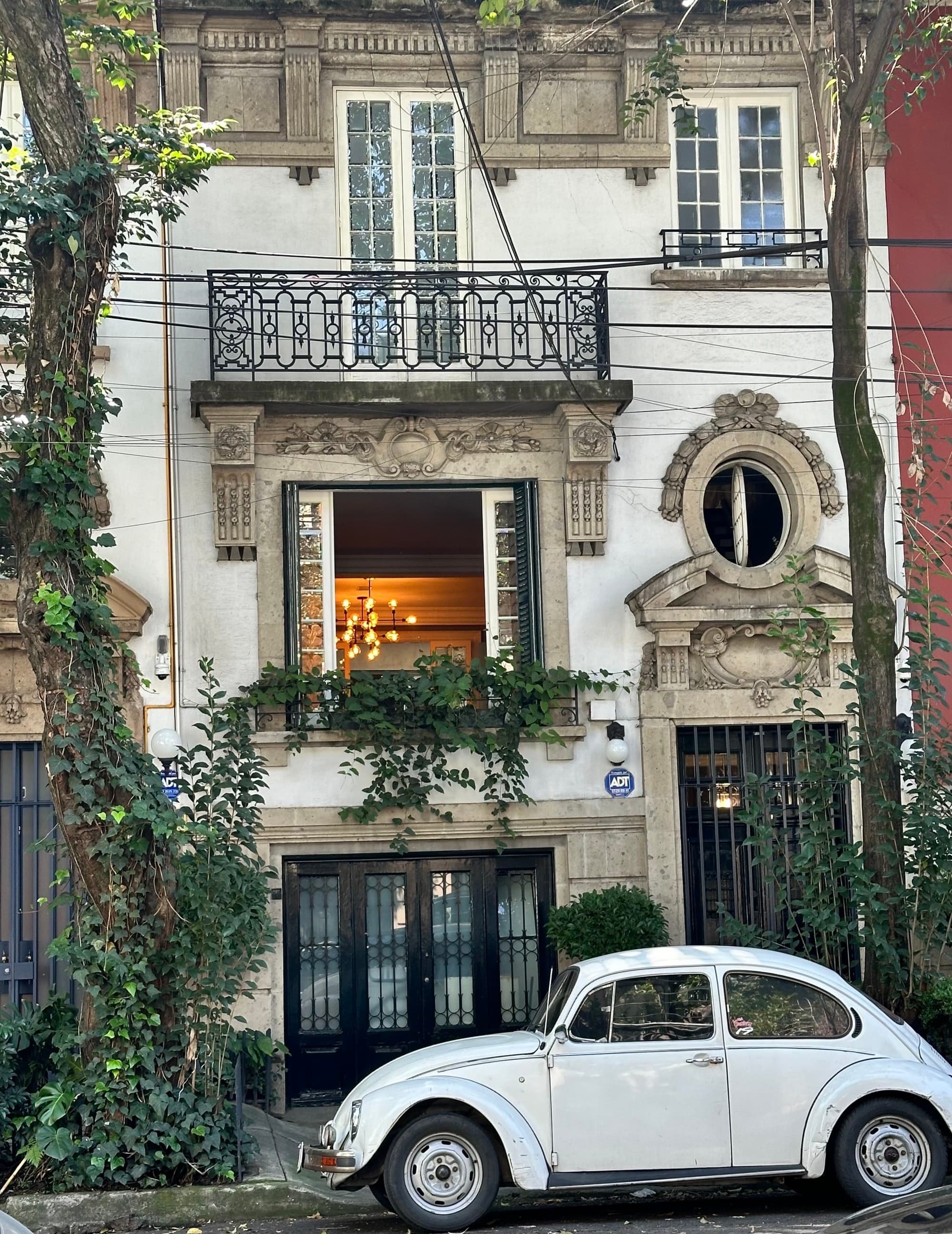 A vintage white Volkswagen Beetle parked in front of an old building with greenery and a lit interior.