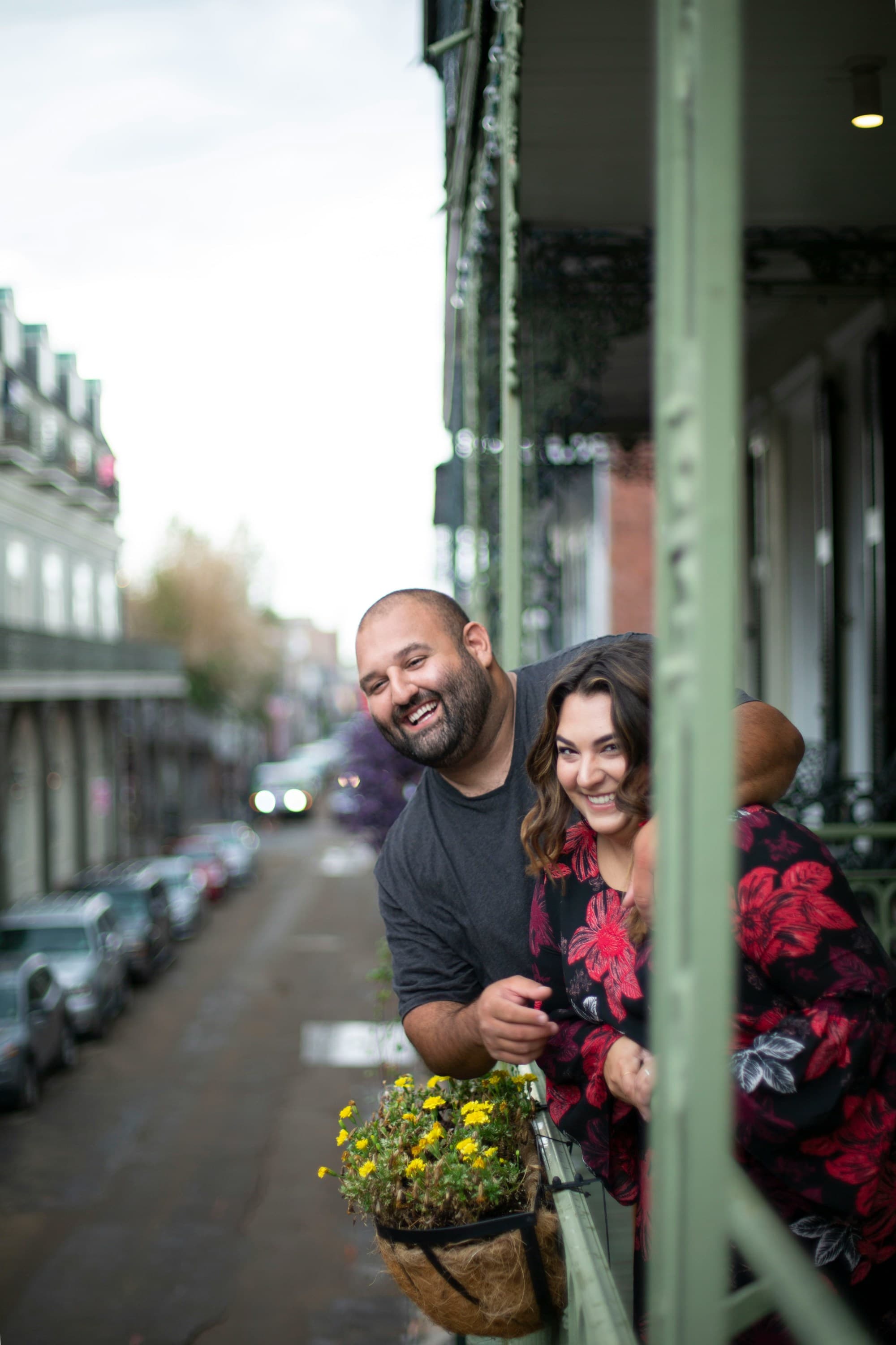 The image shows two individuals standing on a balcony with a street view in the background.