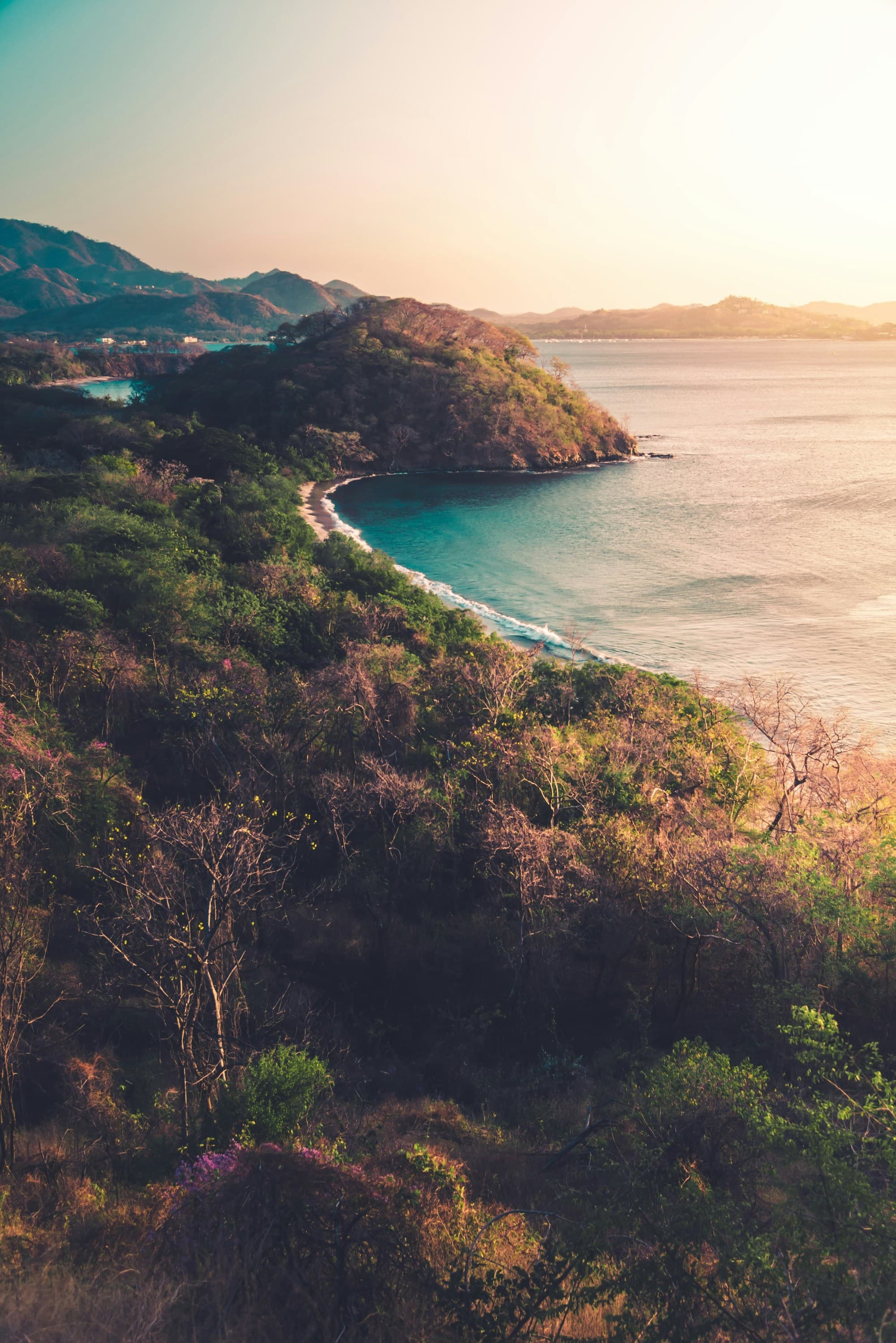 The coastline at sunset with teal water washing calming up onto the tree lined shores with mountains in the distance.