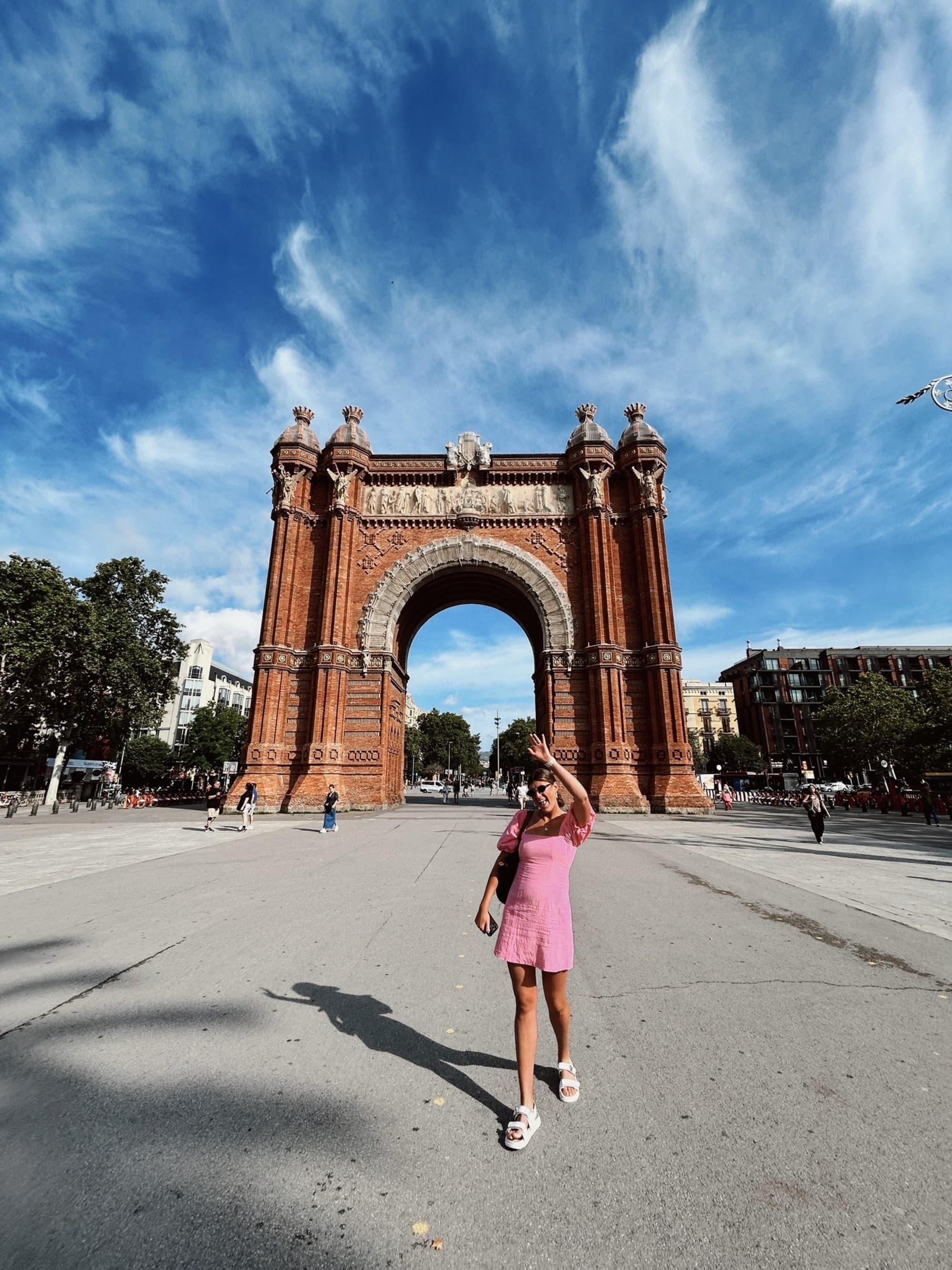 A person standing in front of the Arc de Triomf in Barcelona, capturing the grandeur of the monument under a blue sky with scattered clouds.