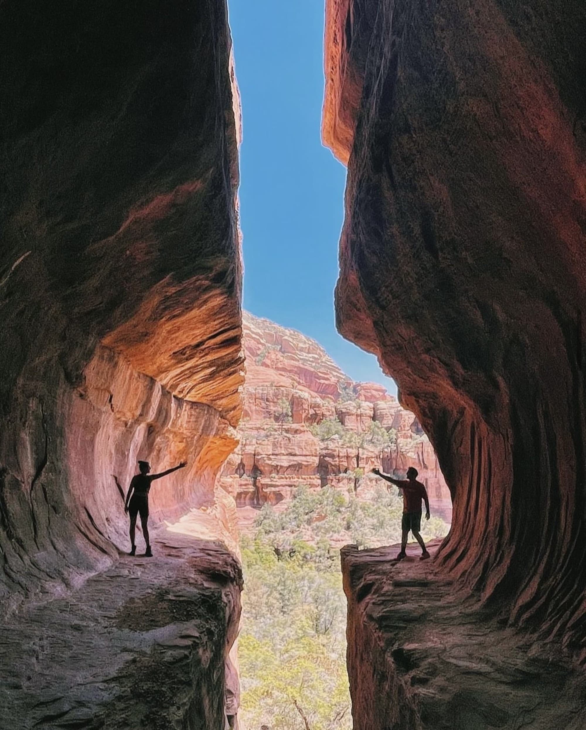 Two silhouettes stand at the edge of a natural divide, bridging the gap between towering cliffs under a vast blue sky.