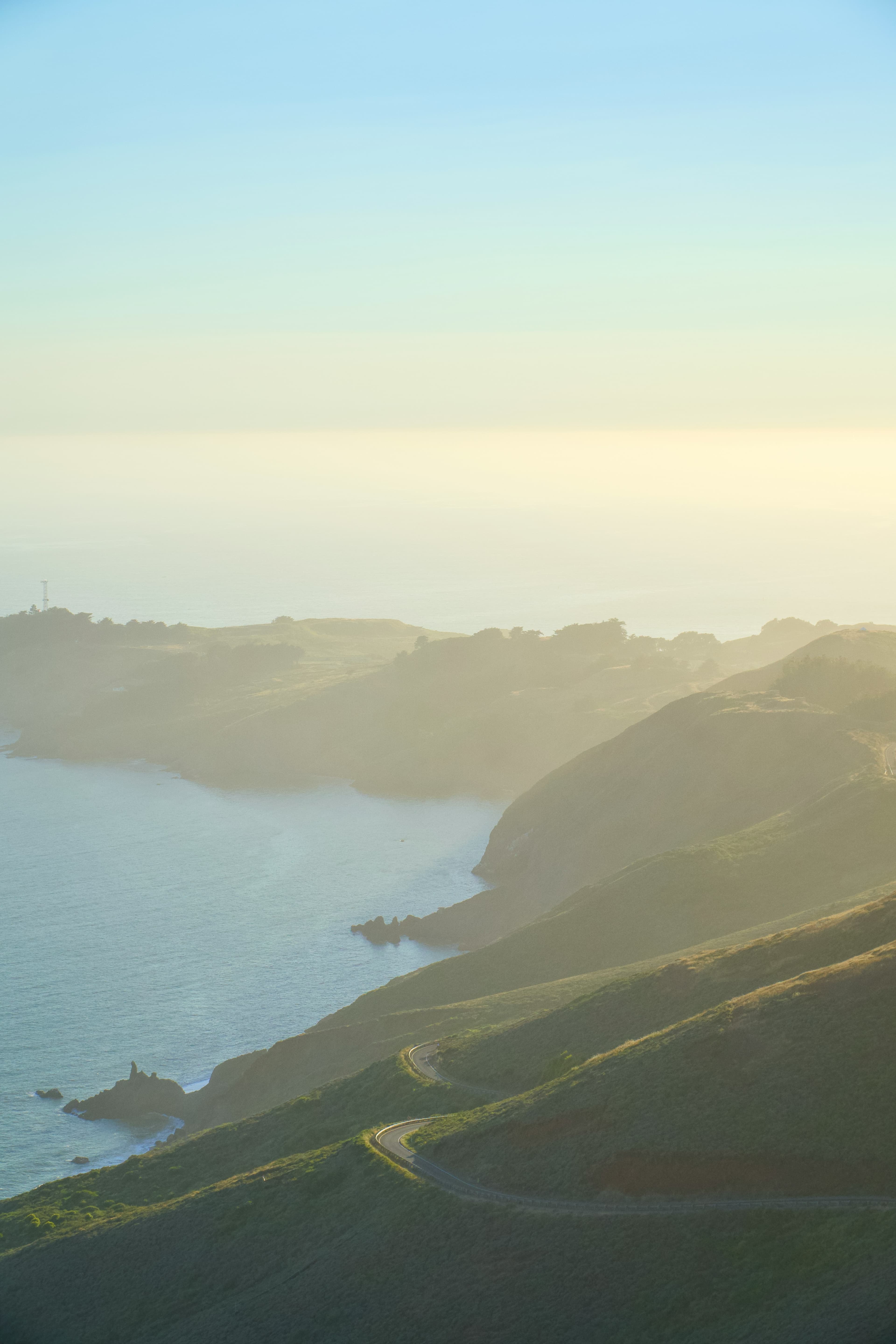 An aerial view of the California coast, with mountains leading to a calm ocean on a hazing day with blue skies.