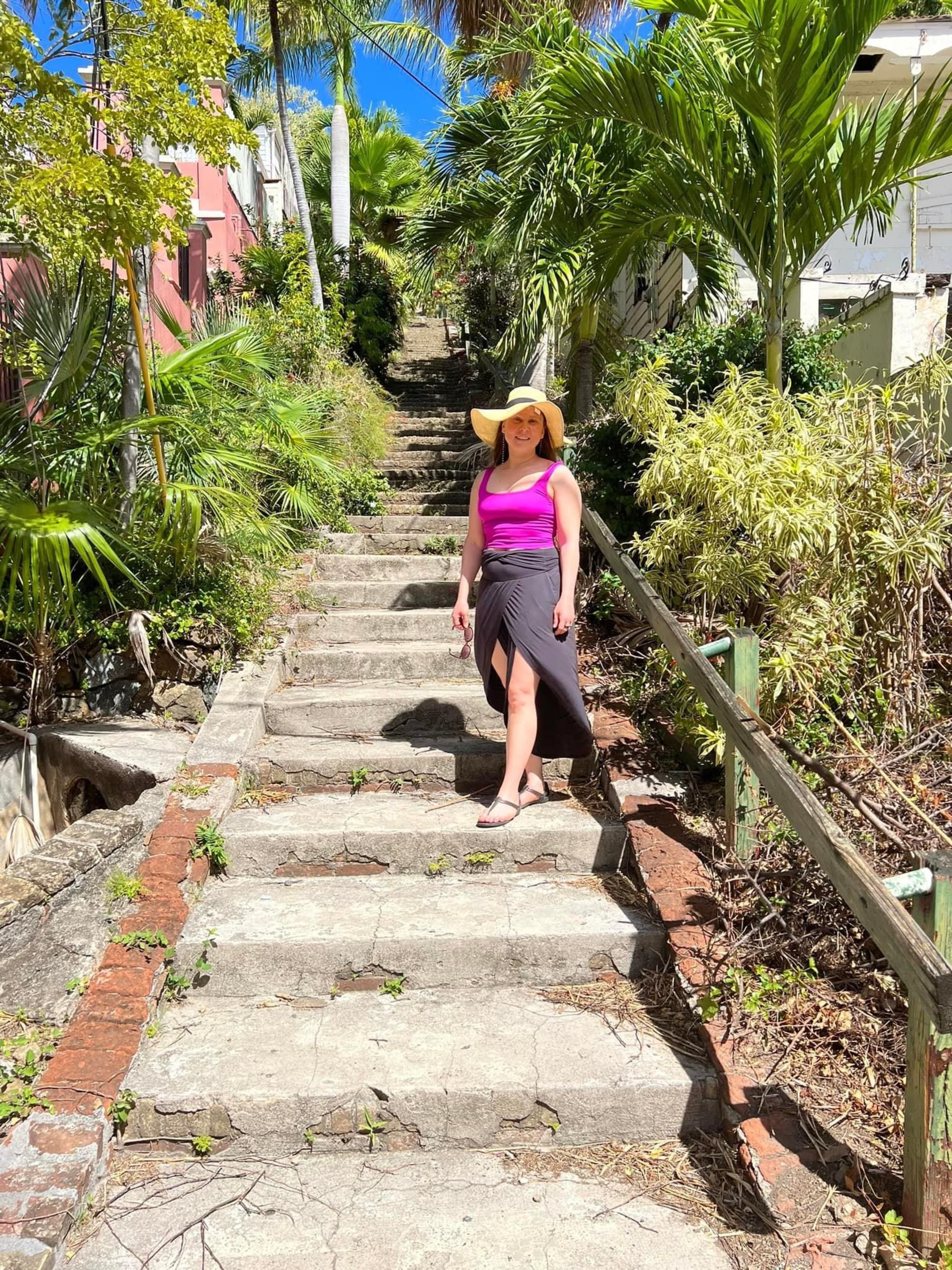 A person in a wide-brimmed sun hat and bright pink top stands on a sunlit stone staircase surrounded by lush tropical greenery.