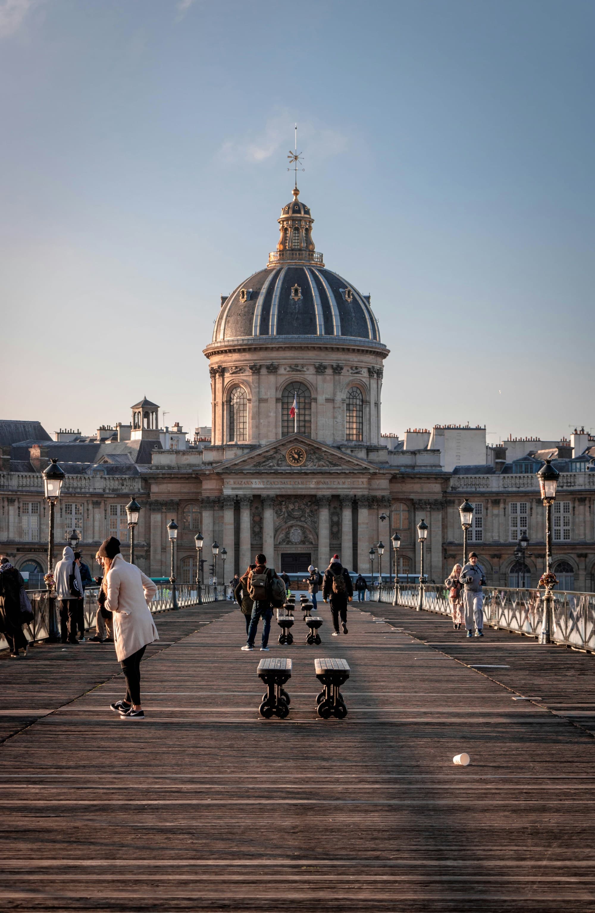 A bridge leading to a grand domed building, bustling with people and street performers under a clear sky.